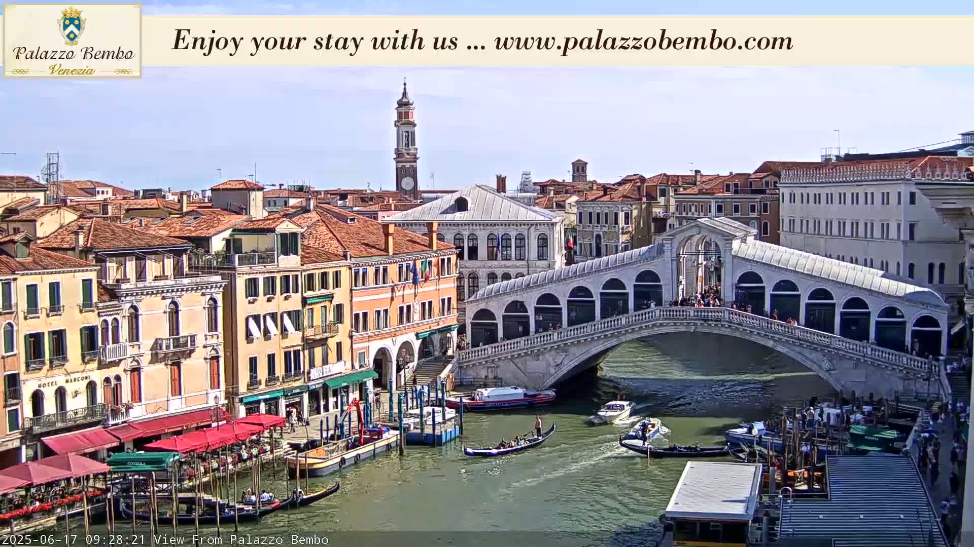 A sunny view of a canal in Venice, with gondolas, boats, and the Rialto Bridge, surrounded by buildings.