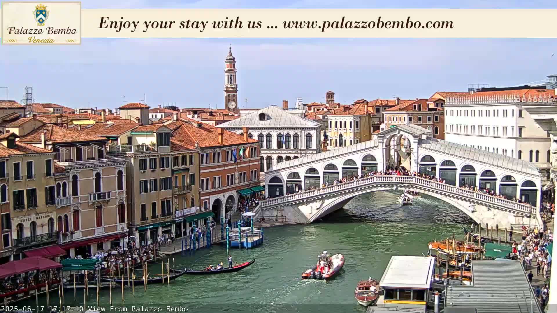A sunny day in Venice shows a canal with gondolas and boats passing under the Rialto Bridge, with many buildings lining the waterway.