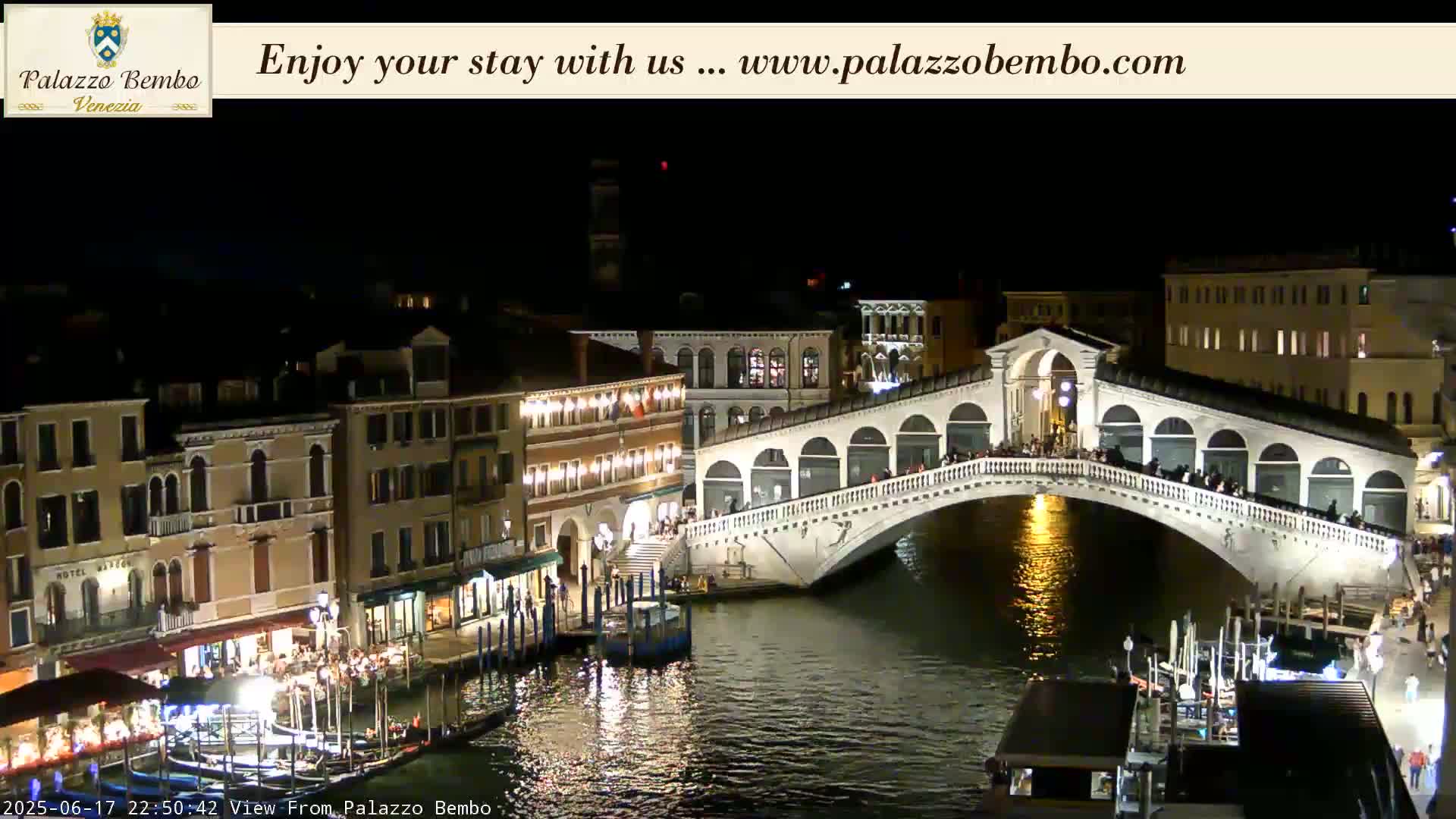 A nighttime view of a canal in Venice, showing a bridge lit up with people walking across it, and buildings along the waterway.