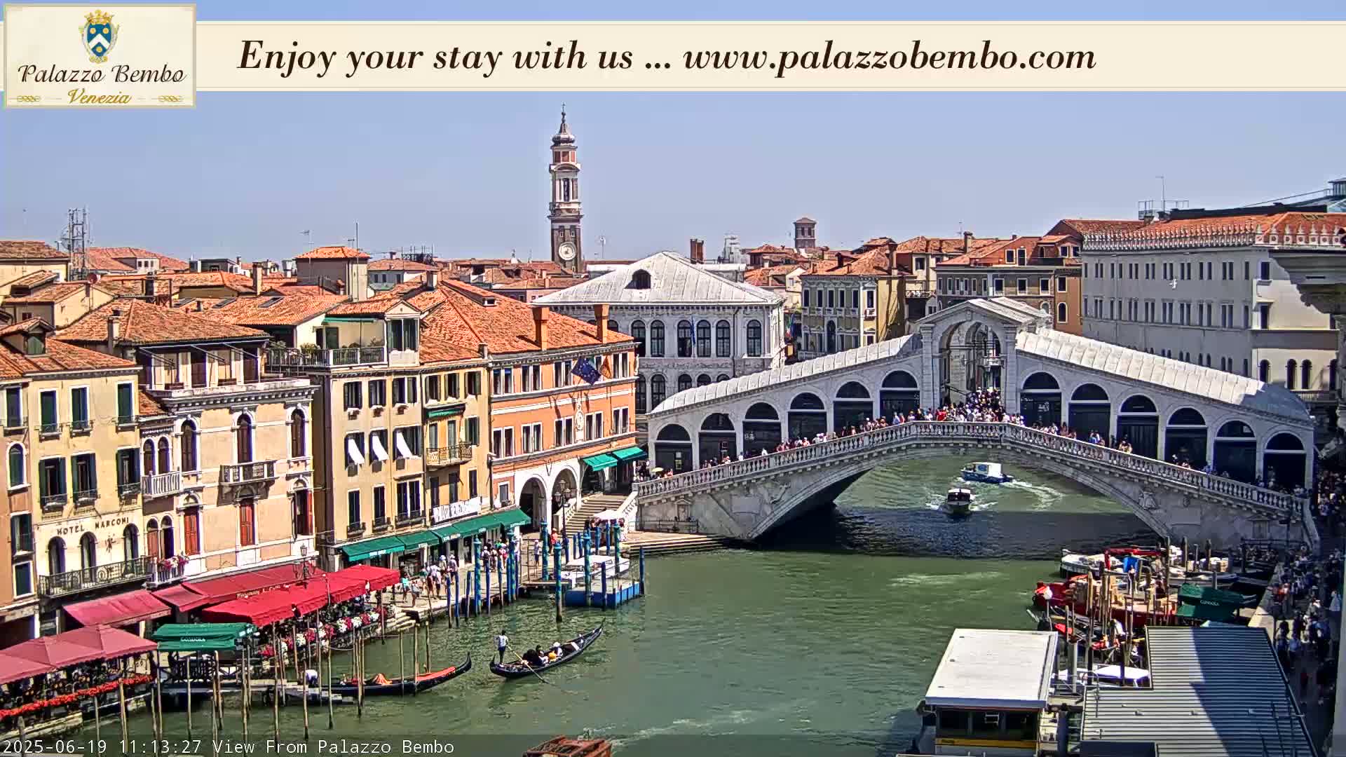 A sunny day in Venice shows a canal with gondolas, boats, and a bridge crowded with people.