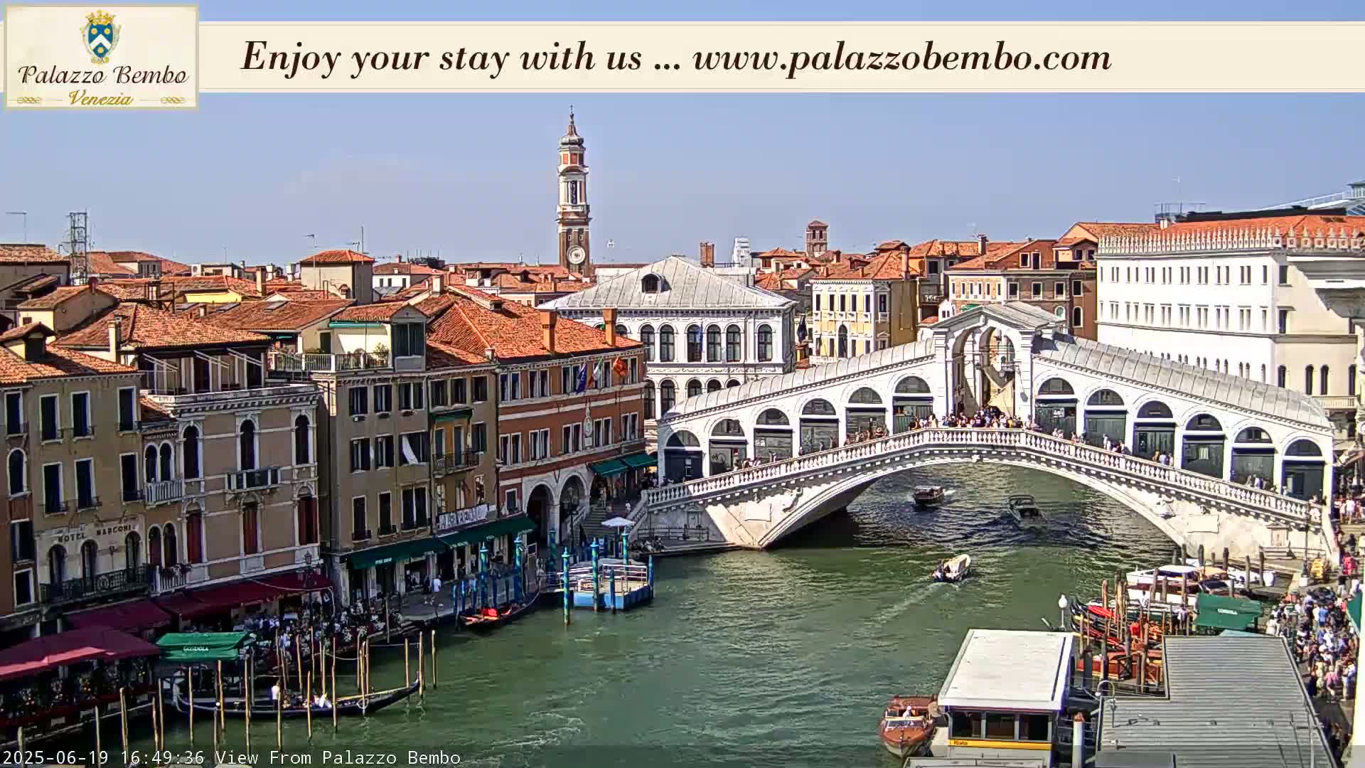 A sunny view of the Rialto Bridge in Venice, with gondolas and boats on the water, and numerous buildings lining the canal.
