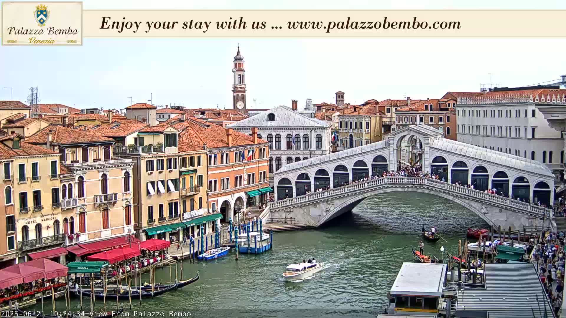 A bridge crowded with people spans a canal in Venice, with colorful buildings lining the waterways under a mostly clear sky.