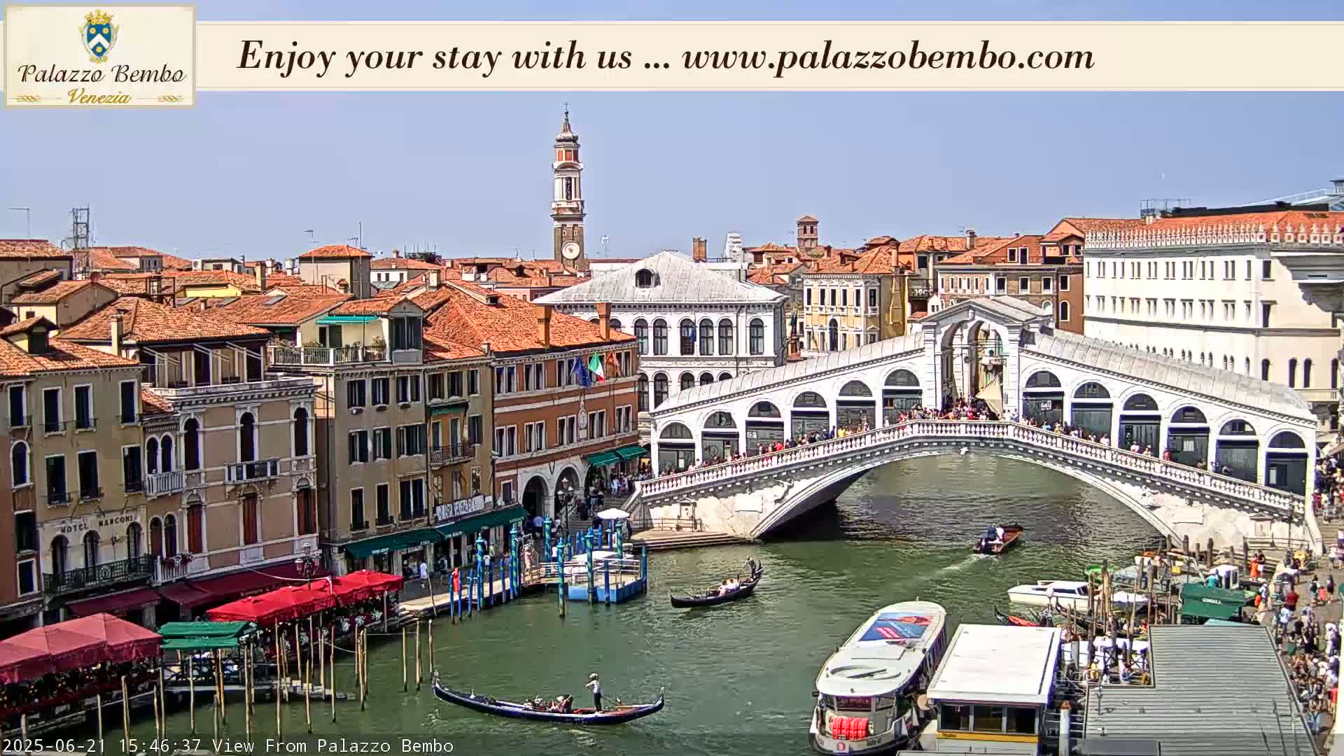 A sunny day in Venice shows a canal with gondolas and a large bridge crowded with people, lined with buildings on either side.