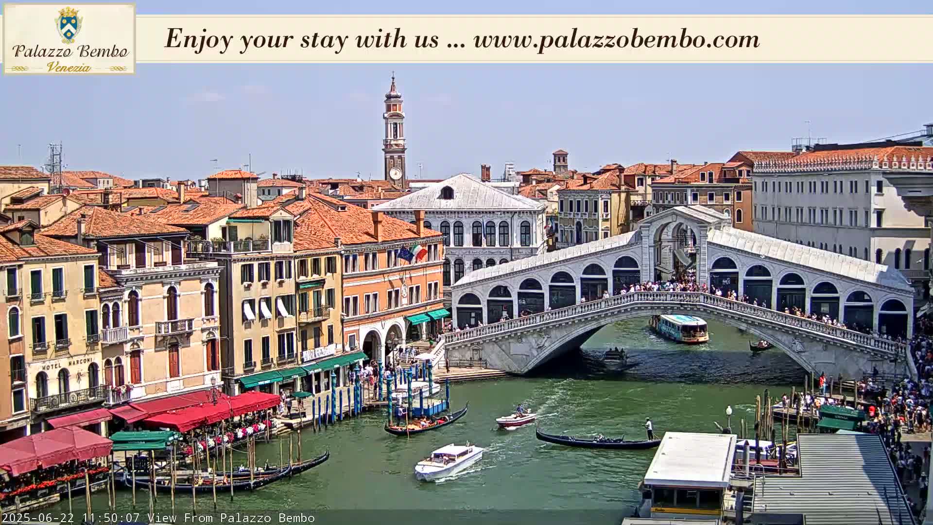 A sunny day in Venice shows a canal with gondolas and boats passing under the Ponte dei Sospiri, with colorful buildings lining the waterway.