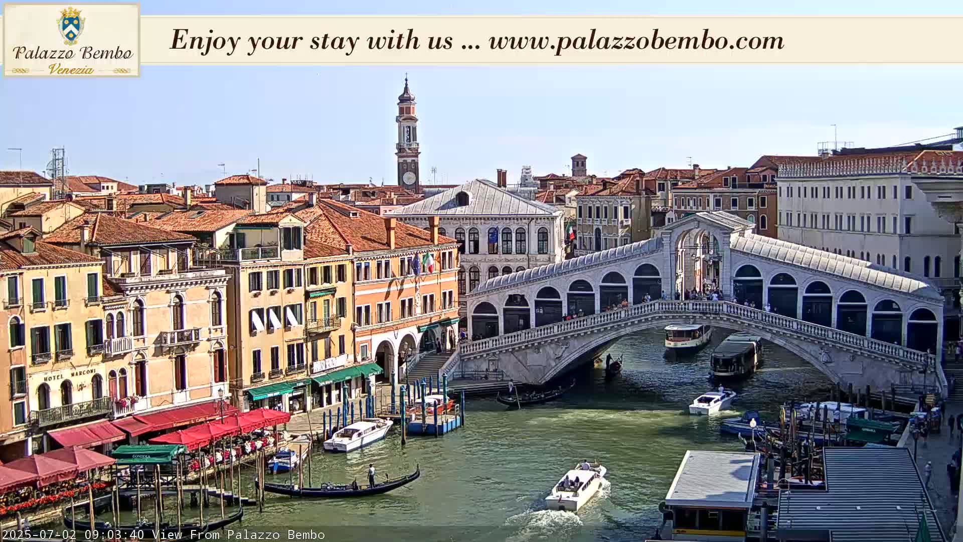 A sunny view of a canal in Venice, Italy, with the Rialto Bridge in the foreground and numerous boats and buildings lining the waterway.
