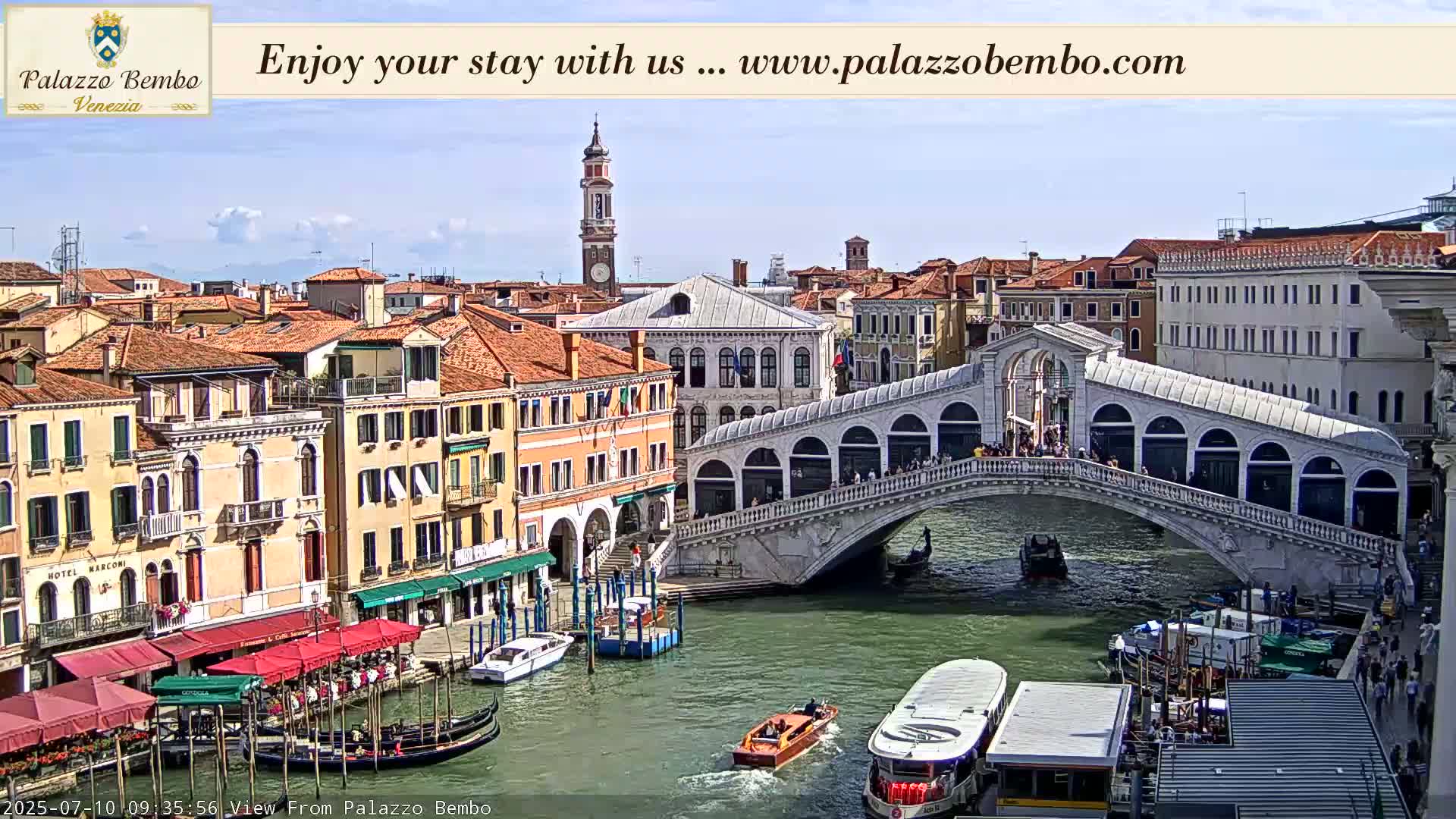 A sunny view of a Venetian canal with gondolas, boats, and the Rialto Bridge, lined with buildings.