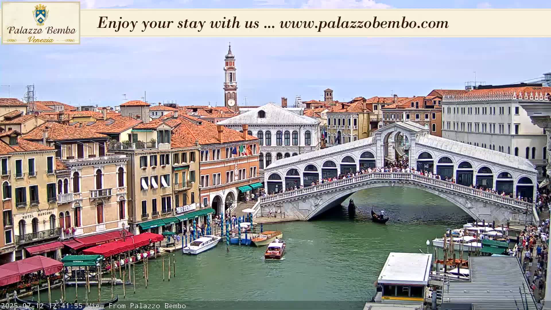 A sunny view of a Venetian canal with gondolas, boats, and the Rialto Bridge, lined with buildings.