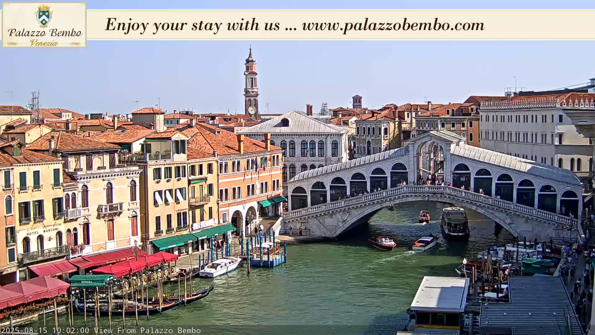 A sunny view of a Venetian canal shows a bridge with people walking across it, boats on the water, and colorful buildings lining the waterway.