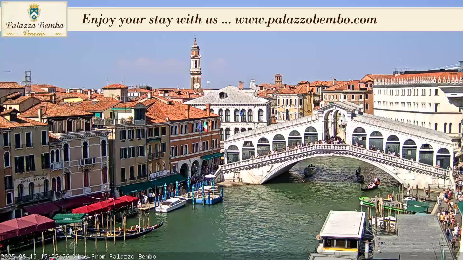 A sunny view of a Venetian canal shows a bridge with people walking across it, boats on the water, and colorful buildings lining the waterway.