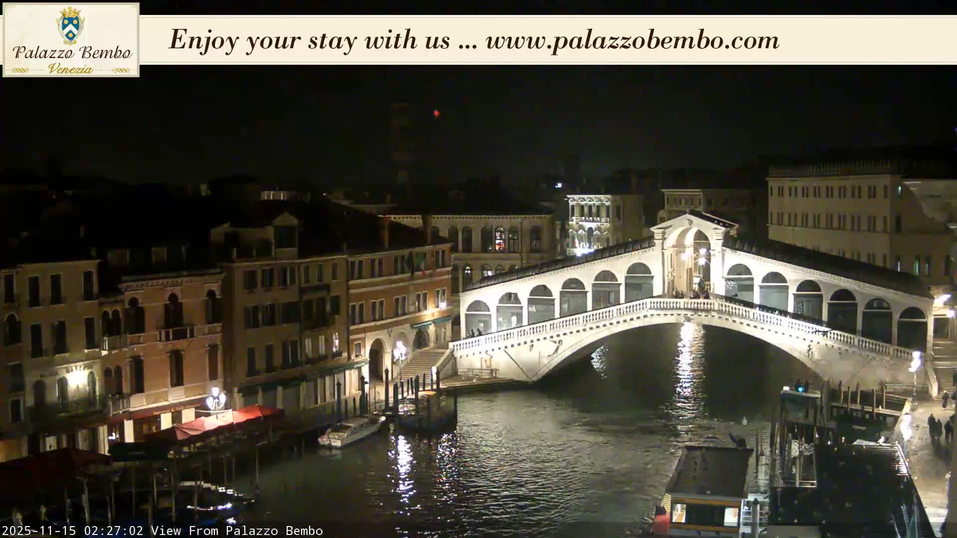 The illuminated Rialto Bridge arches over the Grand Canal in Venice at night, surrounded by historic buildings and boats, with city lights reflecting on the water under clear skies.