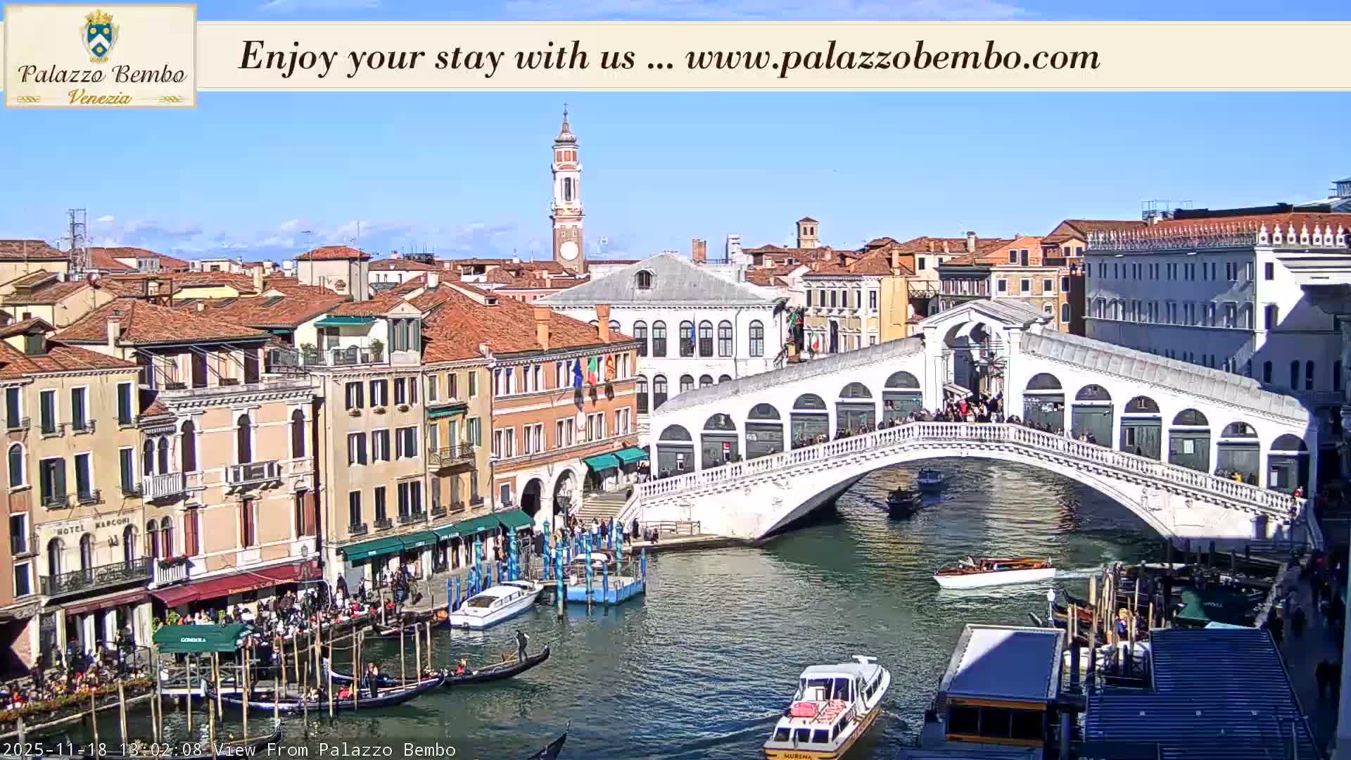 The illuminated Rialto Bridge arches over the Grand Canal in Venice at night, surrounded by historic buildings and boats, with city lights reflecting on the water under clear skies.