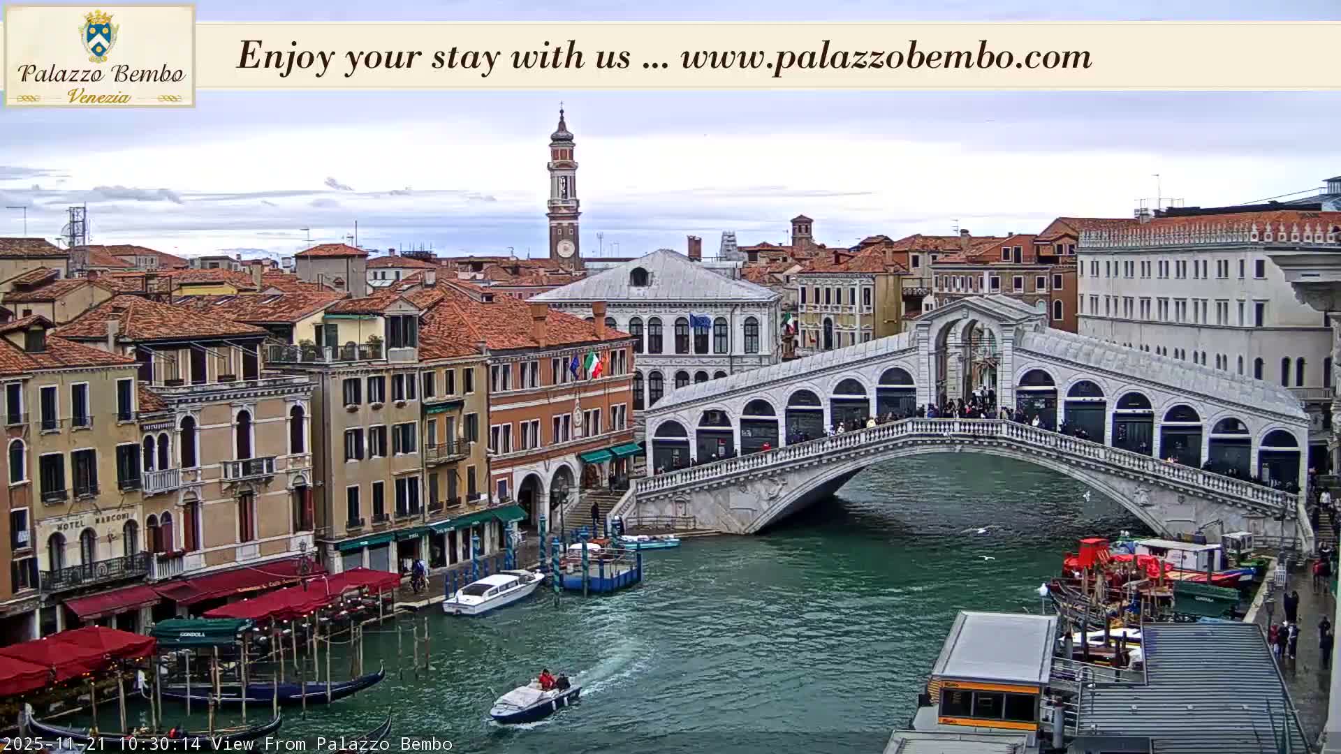 The historic Rialto Bridge, bustling with pedestrians, spans the vibrant Grand Canal where boats navigate among gondolas, all set against a backdrop of classic Venetian architecture under a cloudy sky.