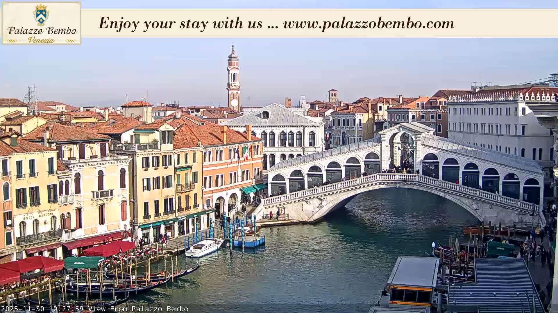 An aerial view of Venice captures the iconic Rialto Bridge arching over the bustling Grand Canal, flanked by historic buildings with red-tiled roofs and numerous gondolas, all under a clear and fair sky.