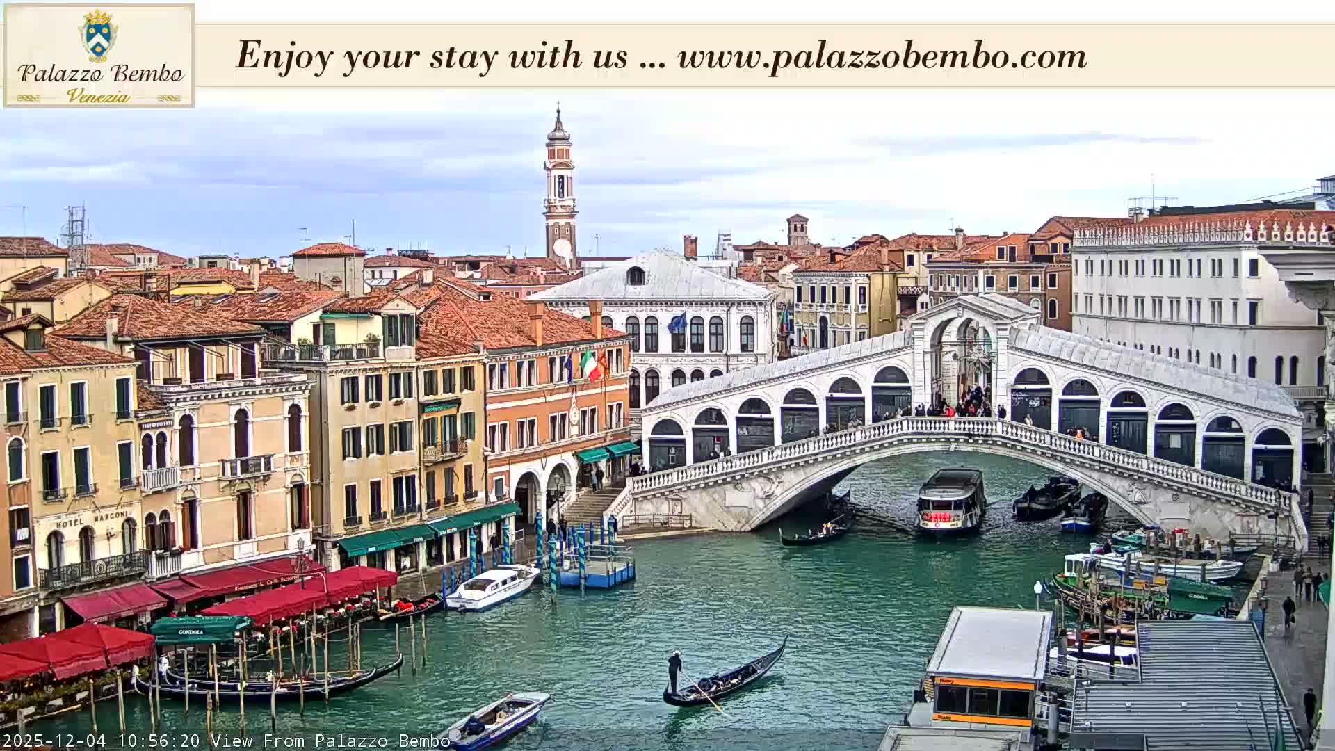 The iconic Rialto Bridge spans the Grand Canal in Venice, flanked by historic buildings and bustling with gondolas and various boats under a partly cloudy sky.