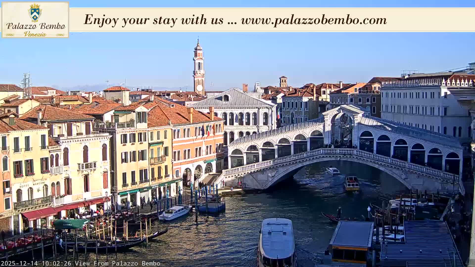The iconic Rialto Bridge spans the Grand Canal in Venice, flanked by historic buildings and bustling with gondolas and various boats under a partly cloudy sky.