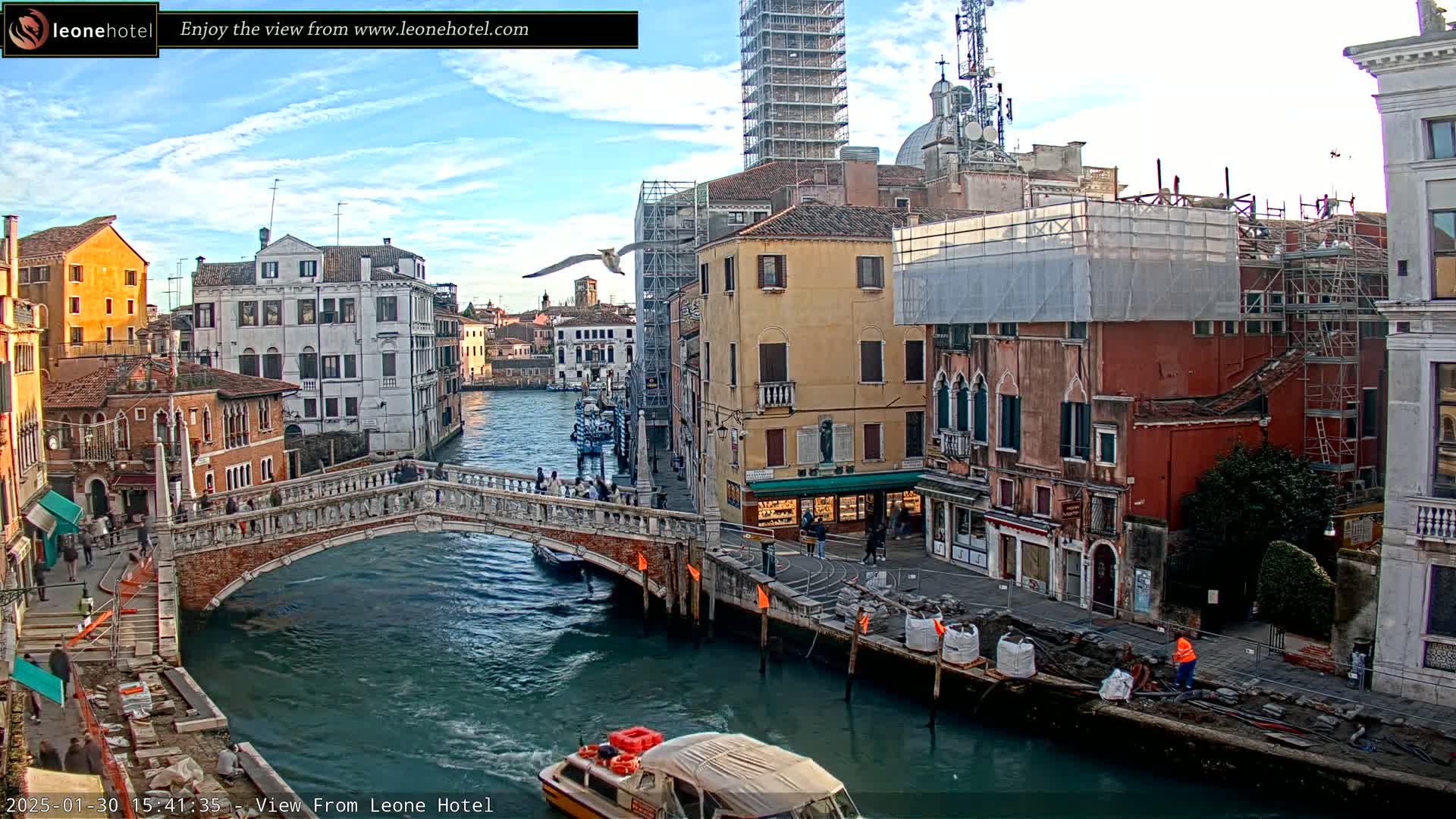 A Venetian canal scene on a partly cloudy day shows a stone bridge, colorful buildings under construction, a boat, and several people walking along the canal.