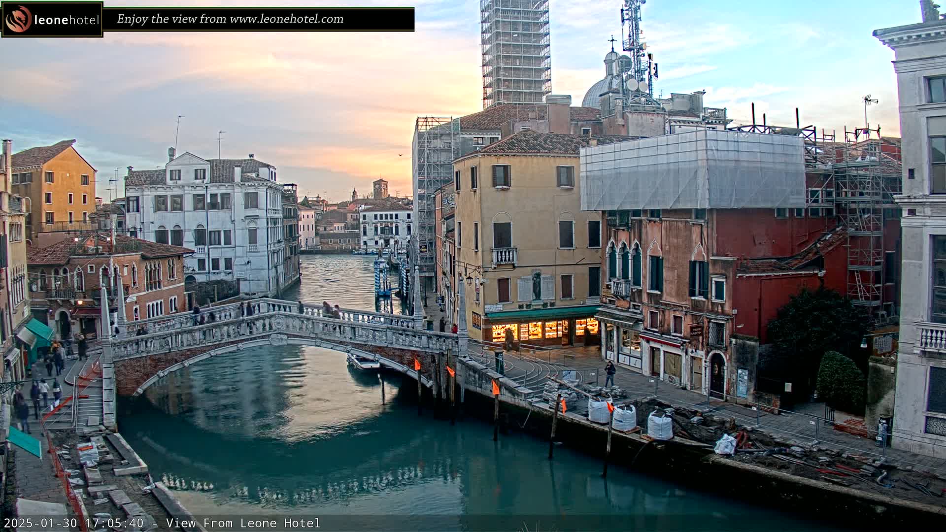 A bridge arches over a canal in Venice, Italy, at sunset, with several buildings under construction or renovation lining the waterway.