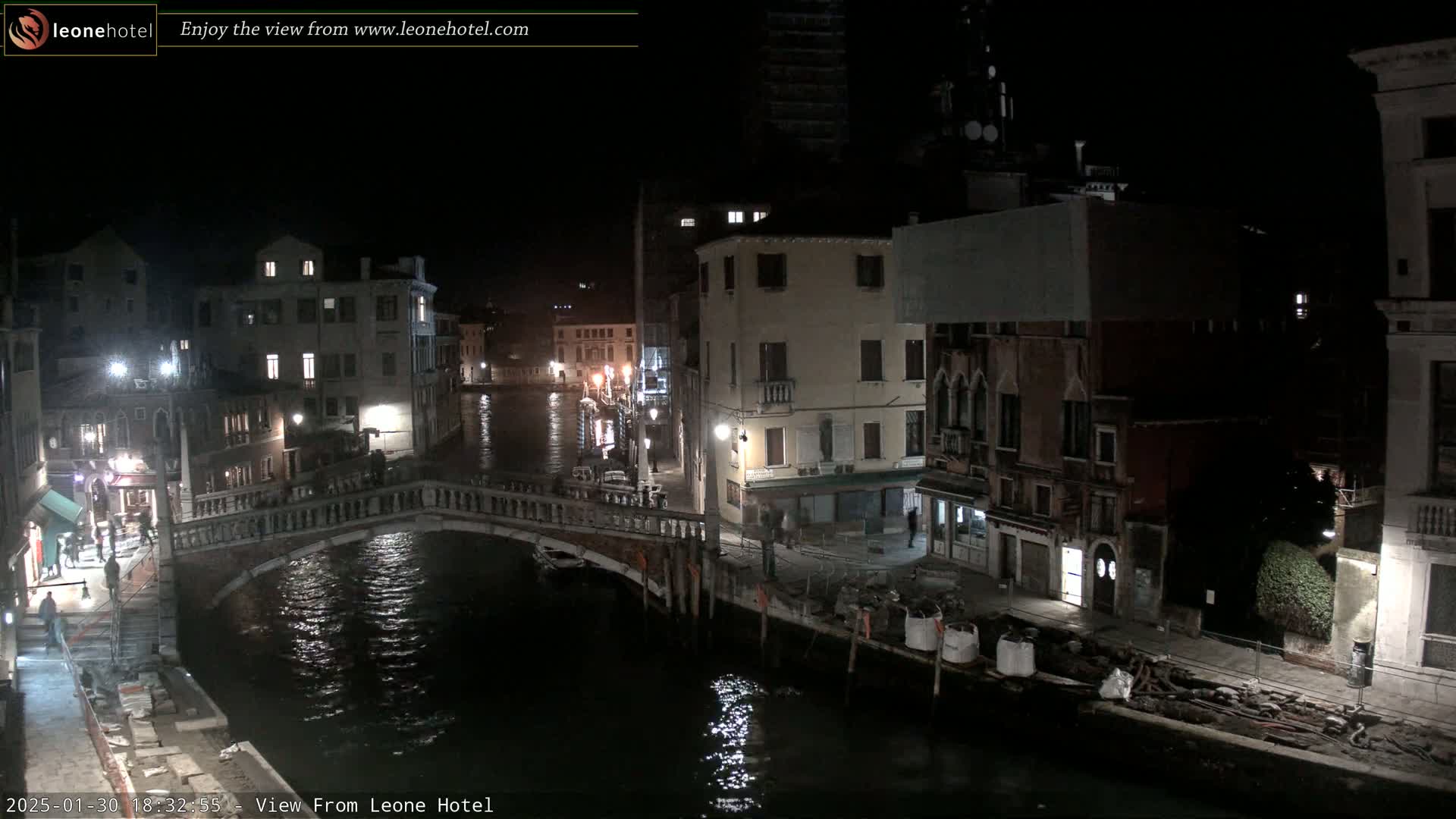 A nighttime view of a canal in Venice, showing a bridge, buildings, and some people walking along the water's edge.
