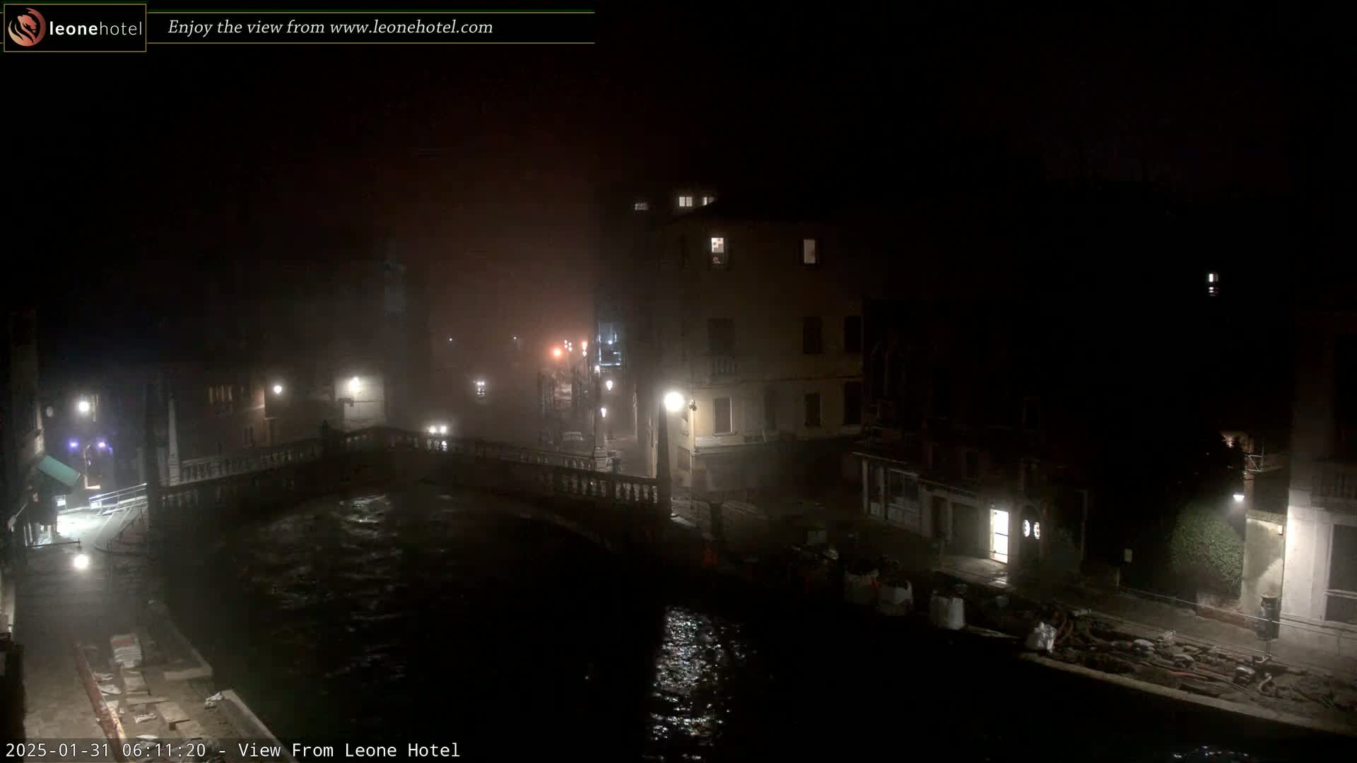 A nighttime view of a foggy canal in Venice, showing bridges, buildings with illuminated windows, and a dark, moving waterway.