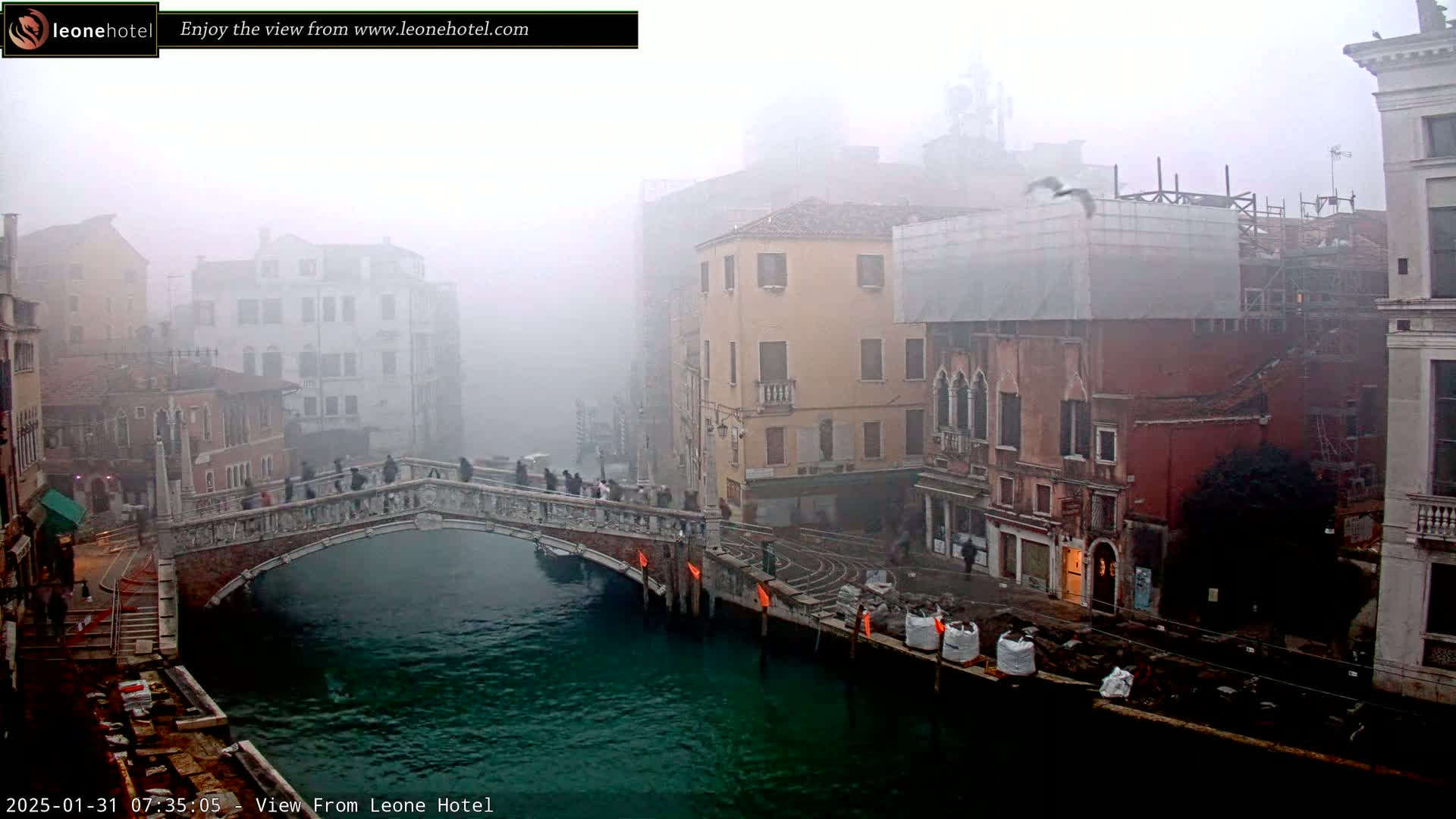 A foggy Venetian canal scene shows a stone bridge with pedestrians, buildings lining the waterway, and construction materials along the canal's edge.