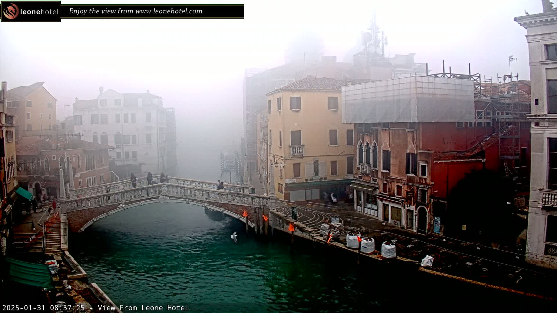 A stone bridge arches over a canal in a foggy Venetian cityscape, with several buildings visible along the waterway.