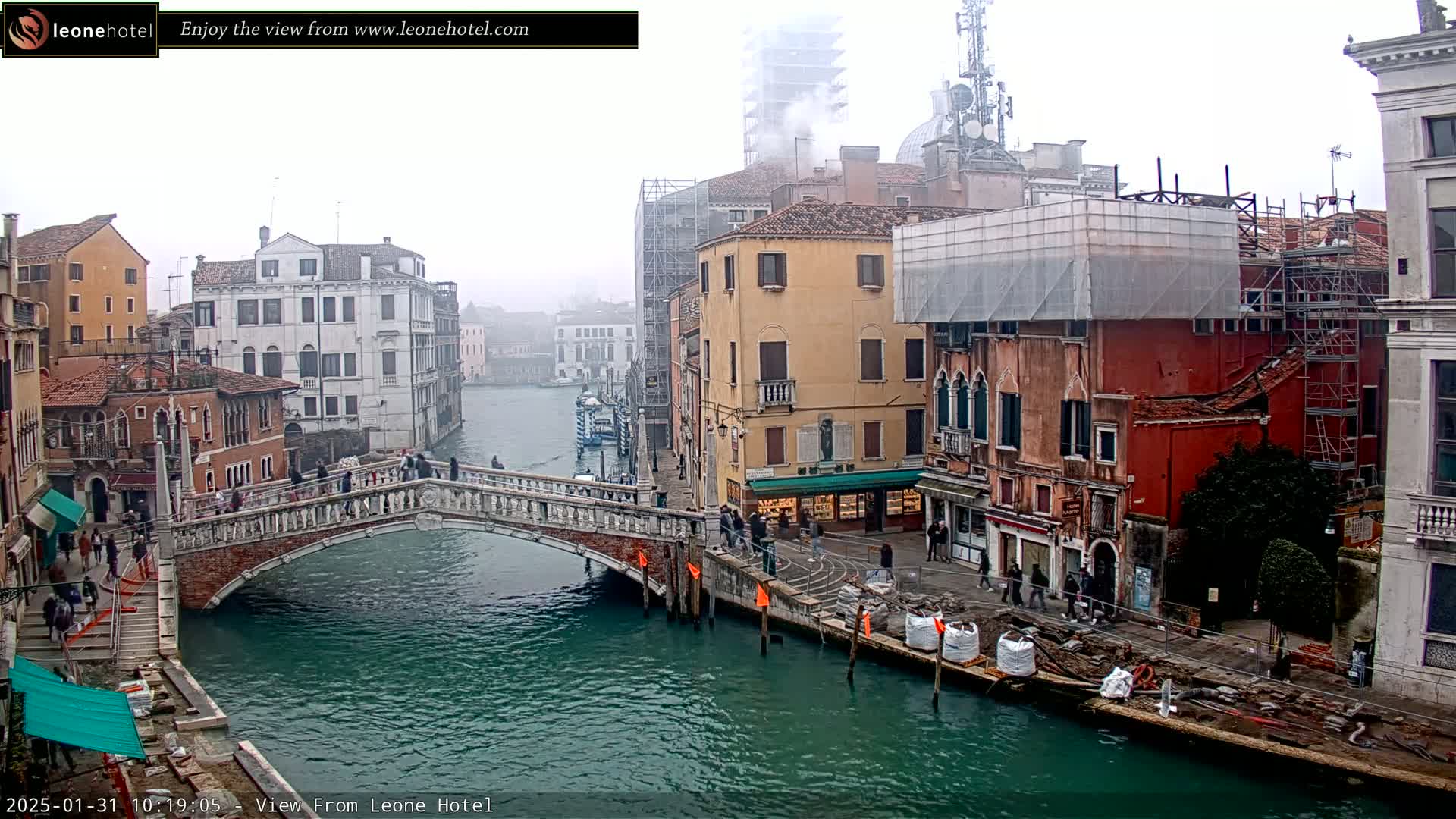 A foggy view of a canal in Venice, Italy, shows several buildings along its banks, a bridge with pedestrians crossing it, and construction materials along the water's edge.