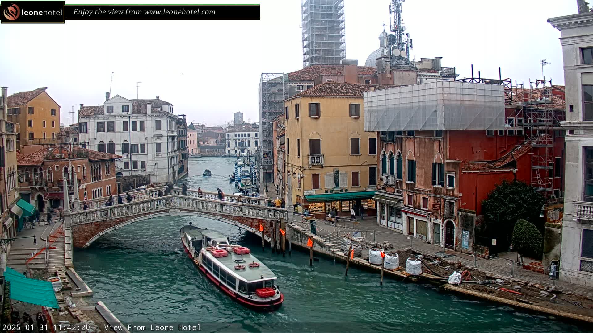 A waterway in Venice, Italy, on an overcast day, shows a bridge with pedestrians, a water taxi, and buildings under construction.