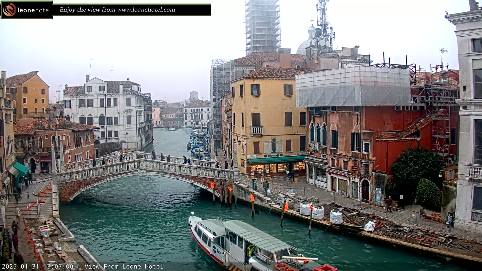 A bridge over a canal in Venice, Italy, on an overcast day, with boats on the water and buildings under construction along the canal's edge.