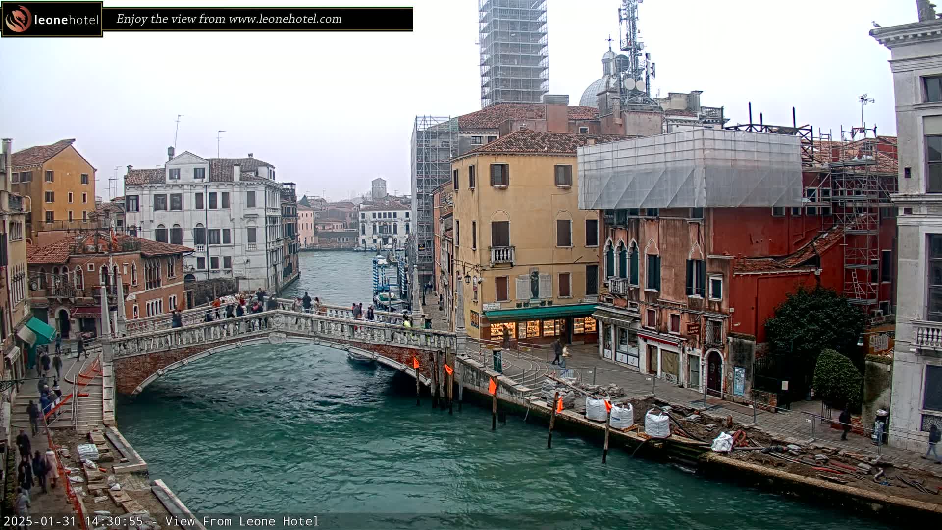 A canal in Venice, Italy, on an overcast day, shows a stone bridge with pedestrians, and several buildings undergoing construction or renovation.