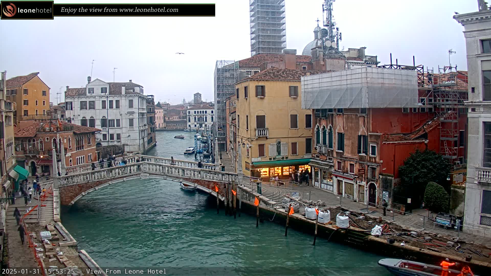 A view of a canal in Venice, Italy, on an overcast day, showing a stone bridge, buildings under construction, and boats on the water.