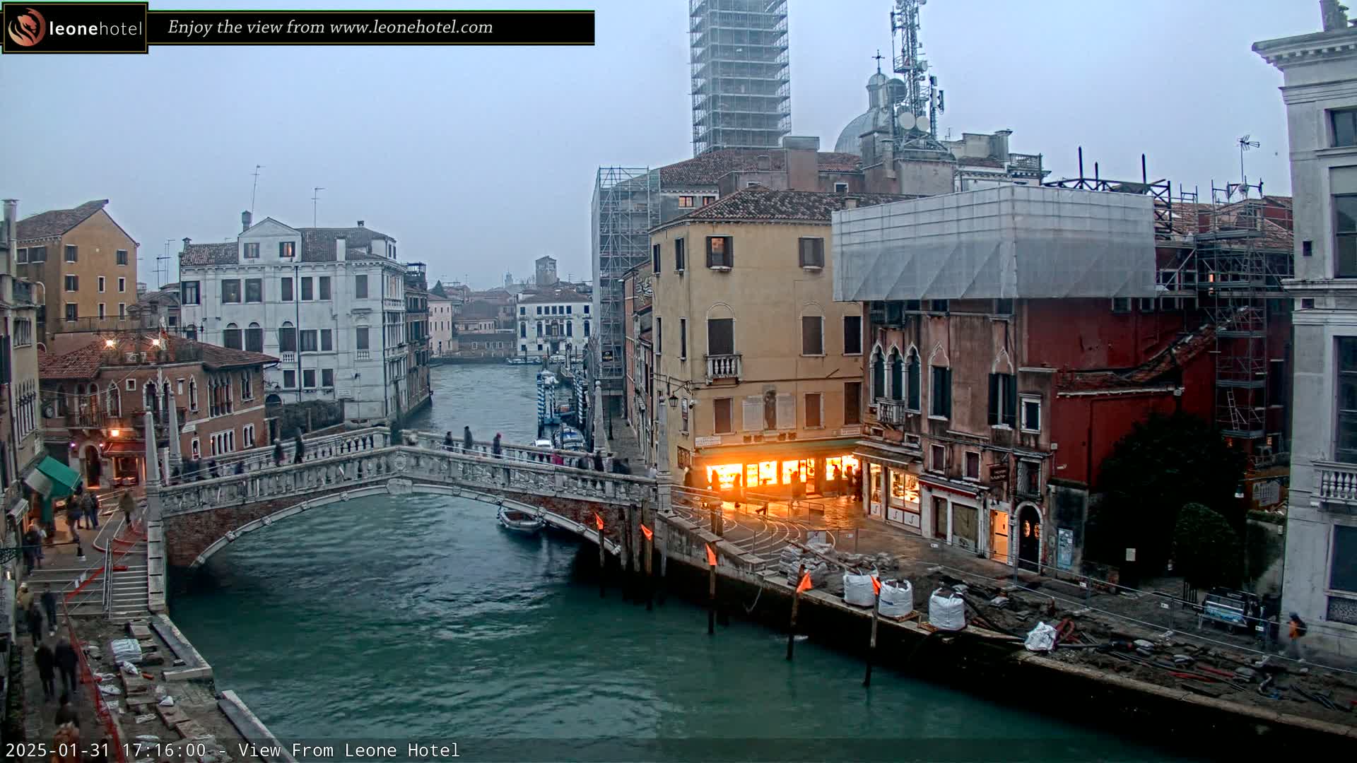 A stone bridge spans a canal in Venice, Italy, under an overcast sky, with several buildings undergoing construction or renovation visible along the waterway.