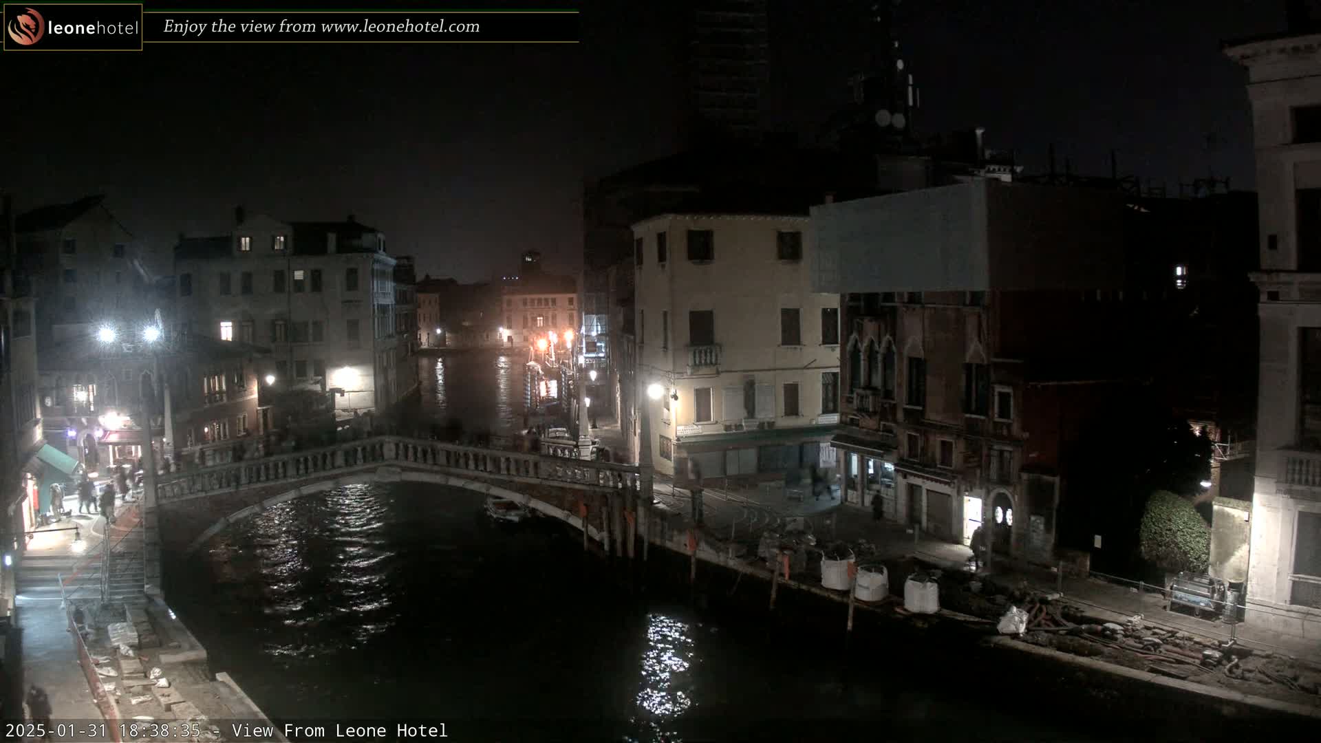 A nighttime view of a canal in Venice, Italy, shows a bridge, buildings, and some people walking across the bridge.