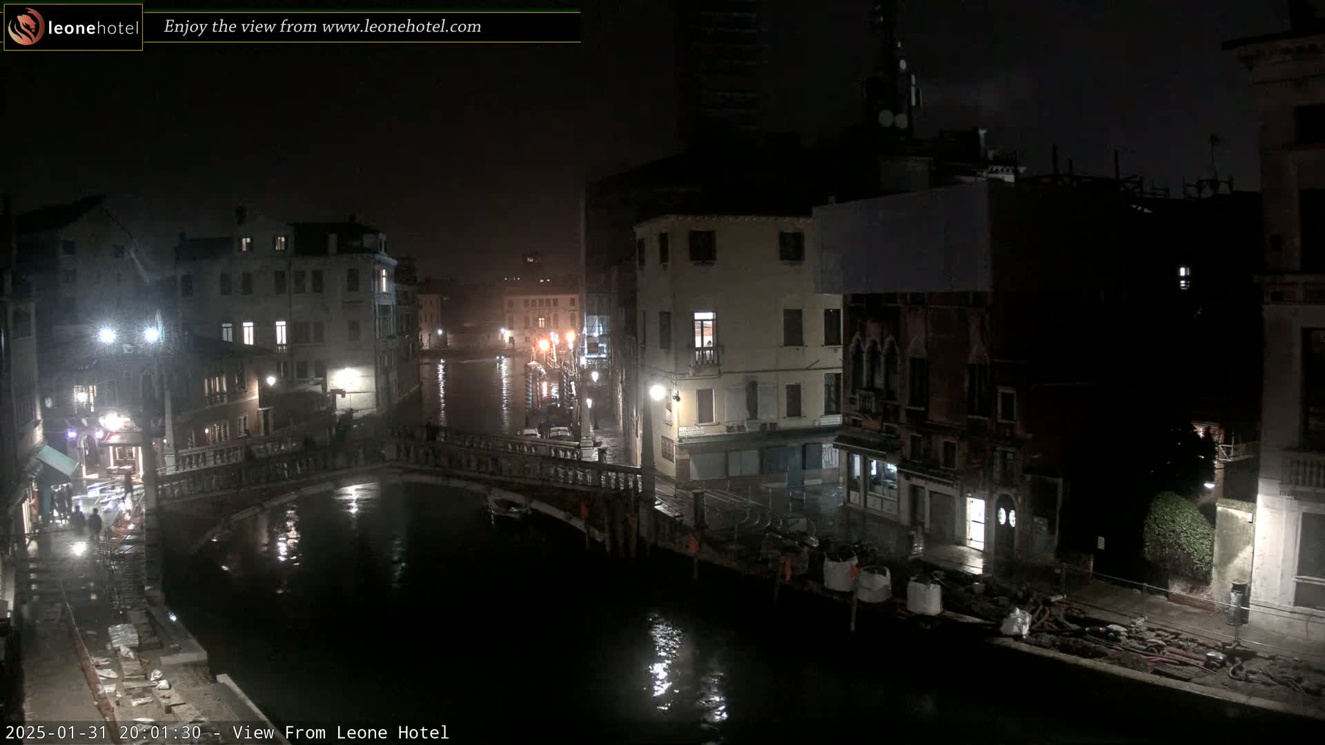 A nighttime view of a canal in Venice, Italy, showing several buildings, a bridge, and wet cobblestones under a dark, possibly rainy sky.