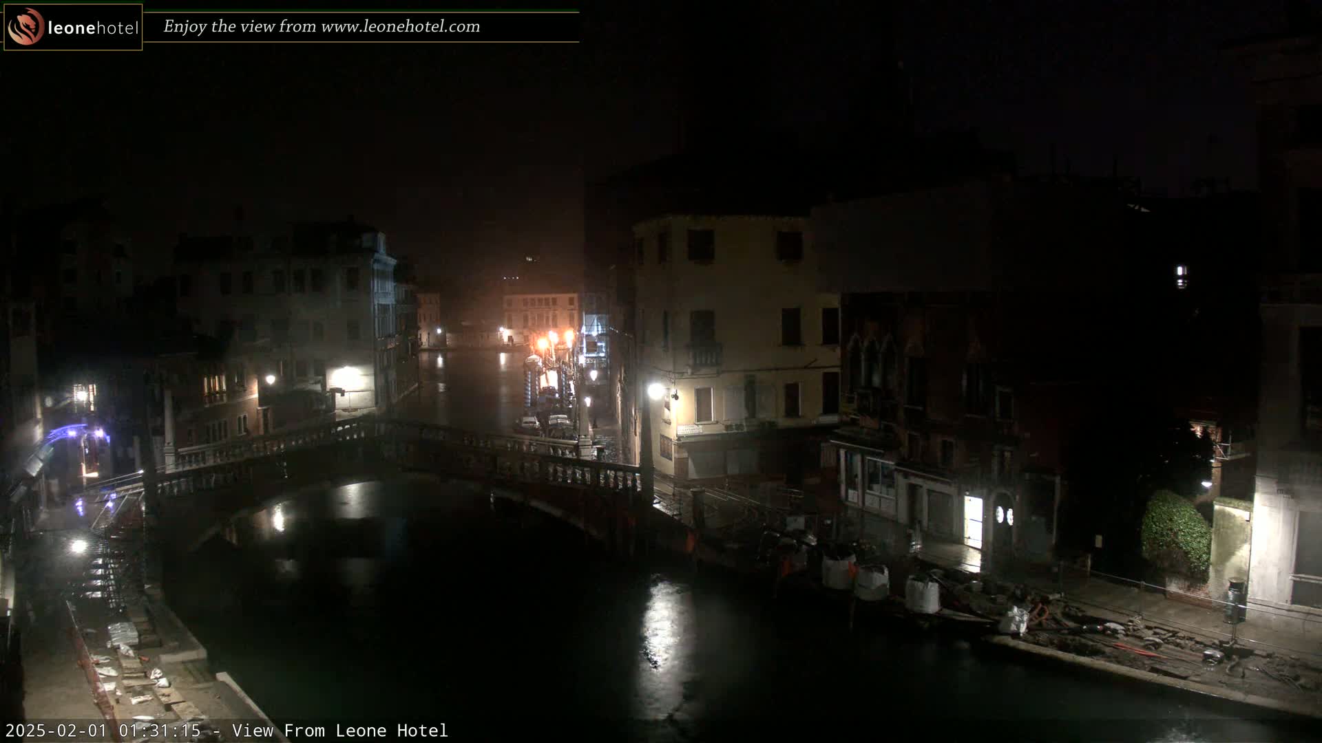 A nighttime view of a canal in Venice, Italy, shows several buildings lining the waterway, with a bridge crossing the canal and some construction or repair work visible along the water's edge.