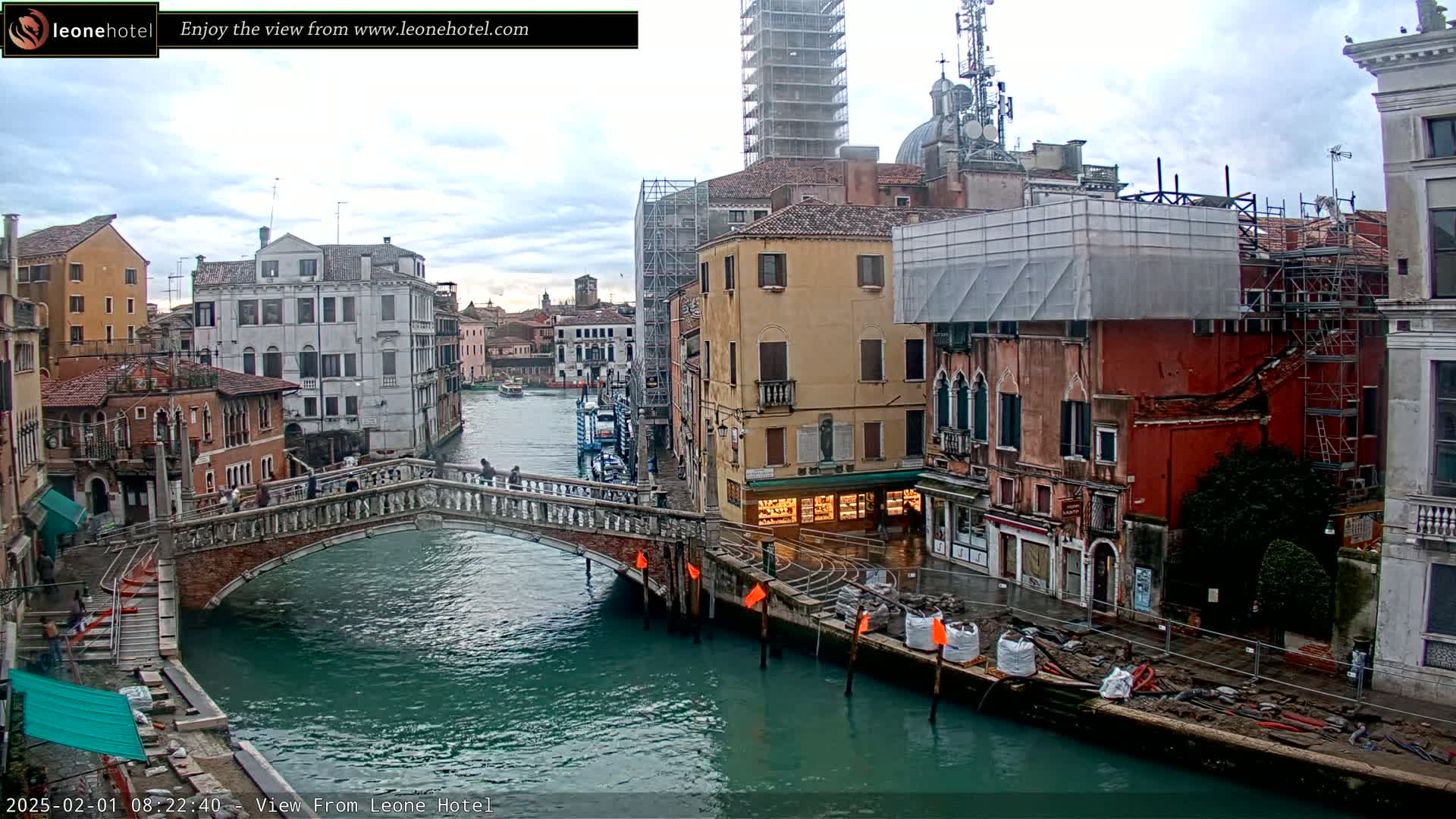 A stone bridge spans a canal in Venice, Italy, under a cloudy sky, with buildings under construction lining the waterway.
