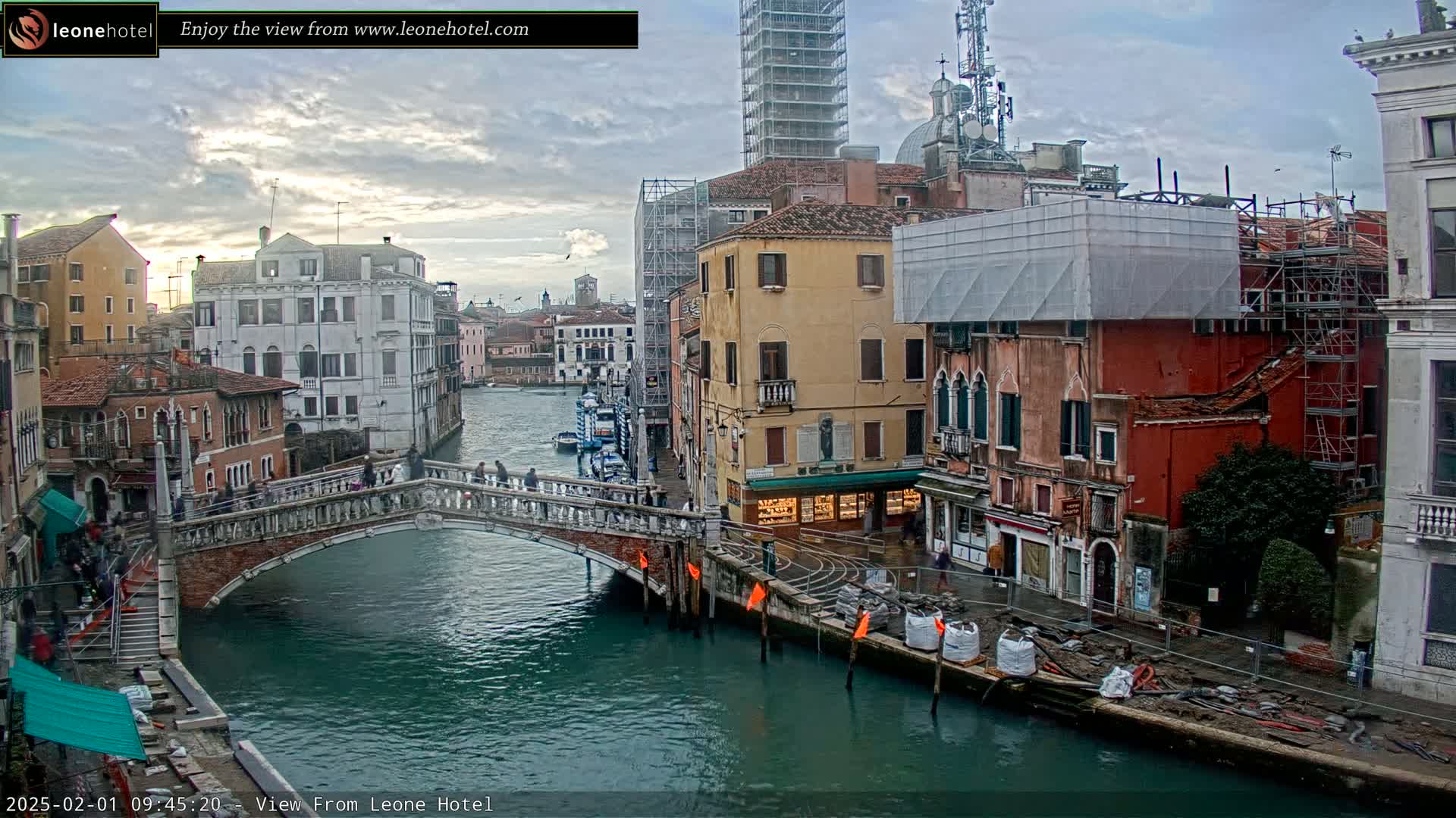 A stone bridge crosses a canal in Venice, Italy, under an overcast sky, with numerous buildings lining the waterway, some undergoing construction.