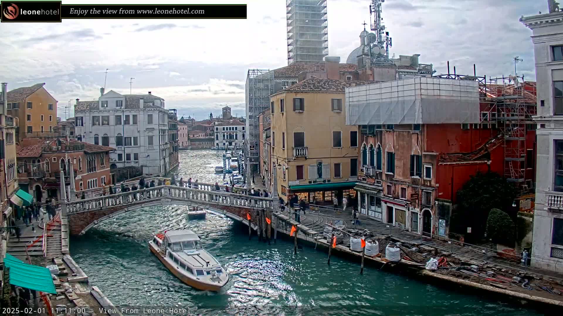 A canal in Venice, Italy, under a cloudy sky, shows a bridge with pedestrians, boats on the water, and buildings undergoing construction.