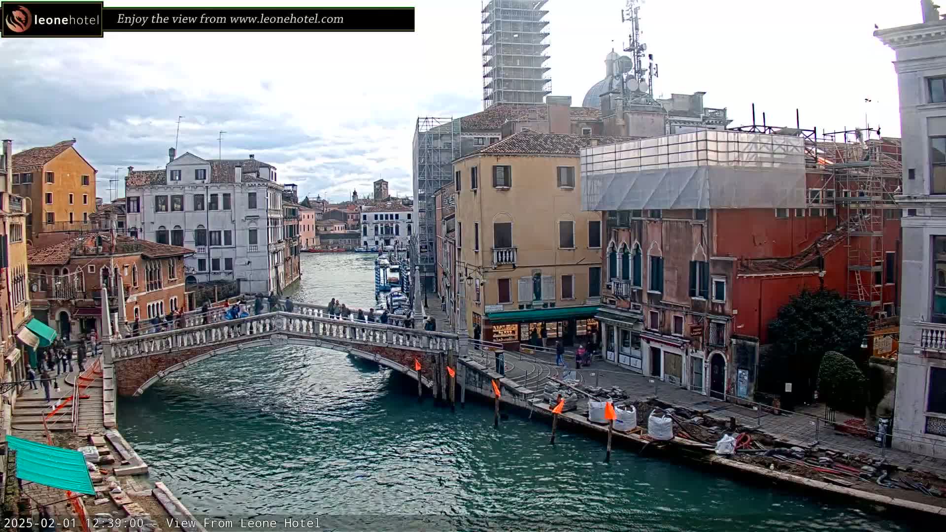 A canal in Venice, Italy, with overcast skies, shows several buildings, a bridge with pedestrians, and ongoing construction along the waterway.