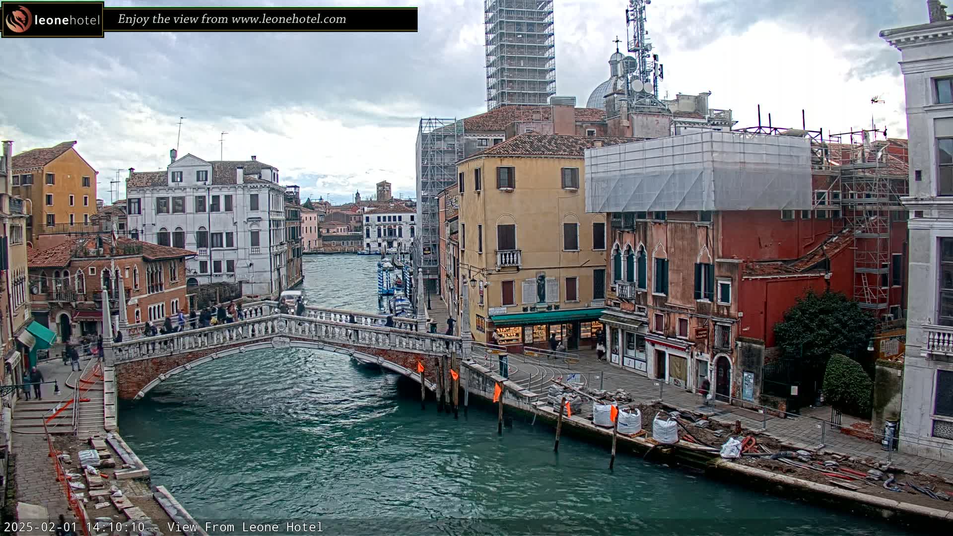 A canal in Venice, Italy, under a cloudy sky, shows several buildings, a bridge with pedestrians, and construction materials along the canal's edge.
