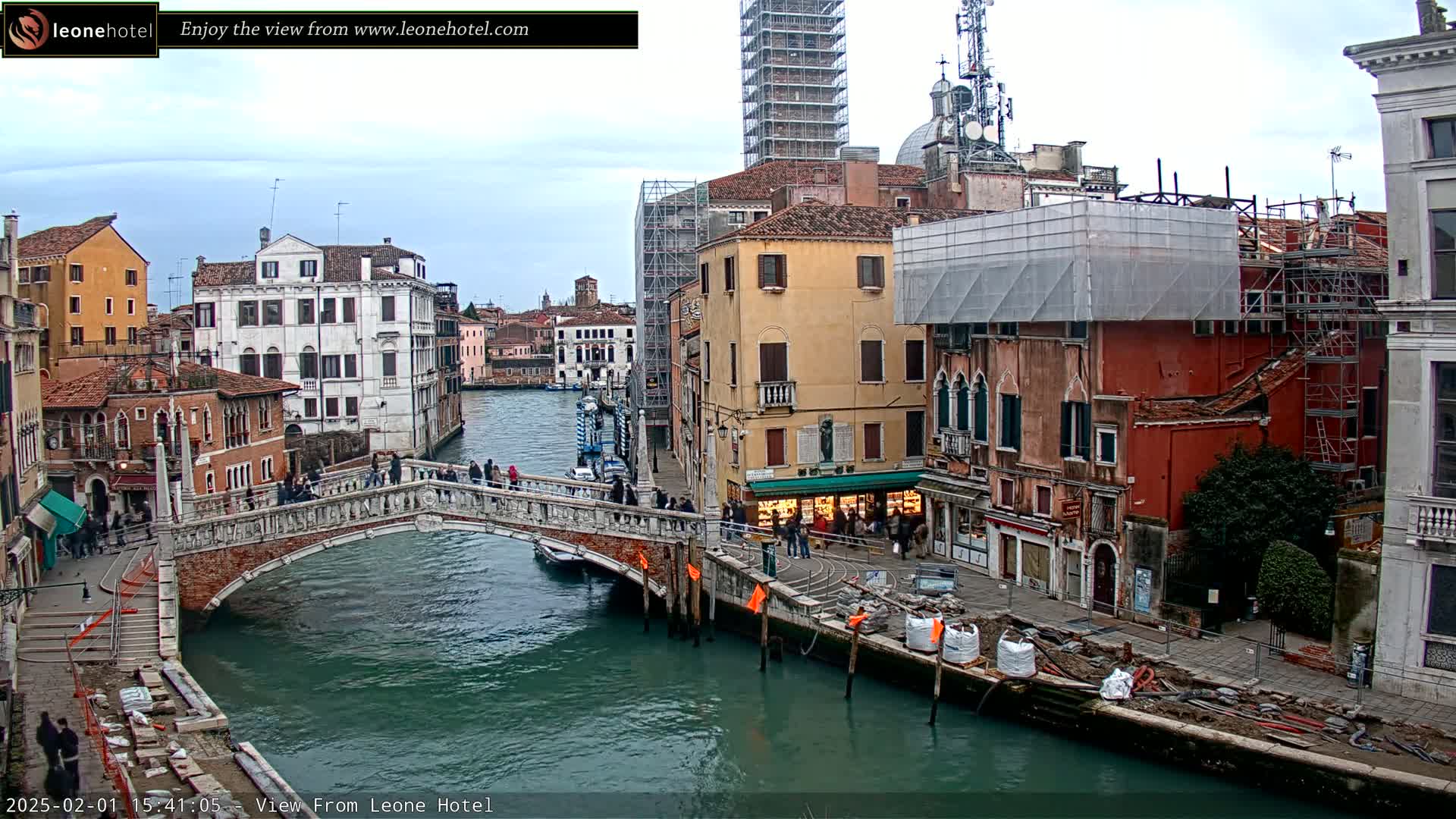 A stone bridge spans a canal in Venice, Italy, under an overcast sky, with several buildings undergoing construction or renovation along the waterway.
