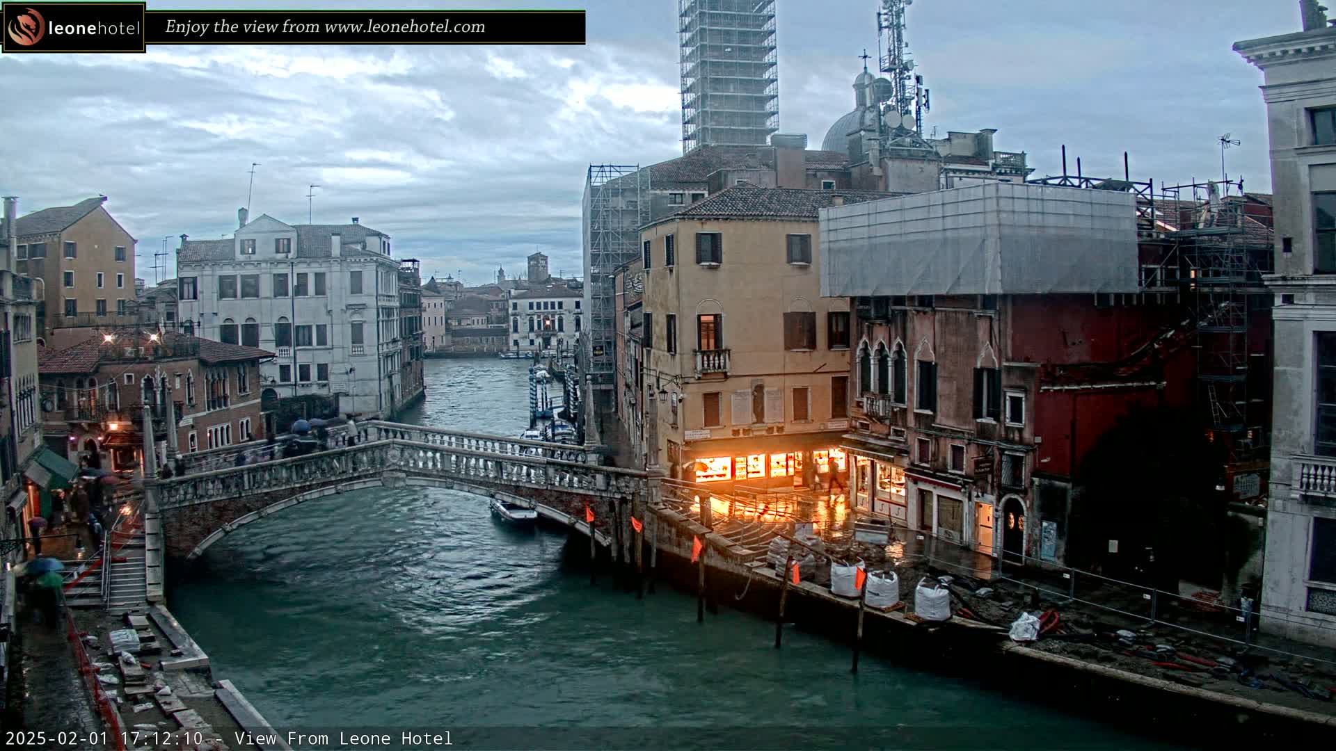 A Venetian canal scene under an overcast sky shows a stone bridge with pedestrians, boats on the water, and several buildings along the canal, some under construction.
