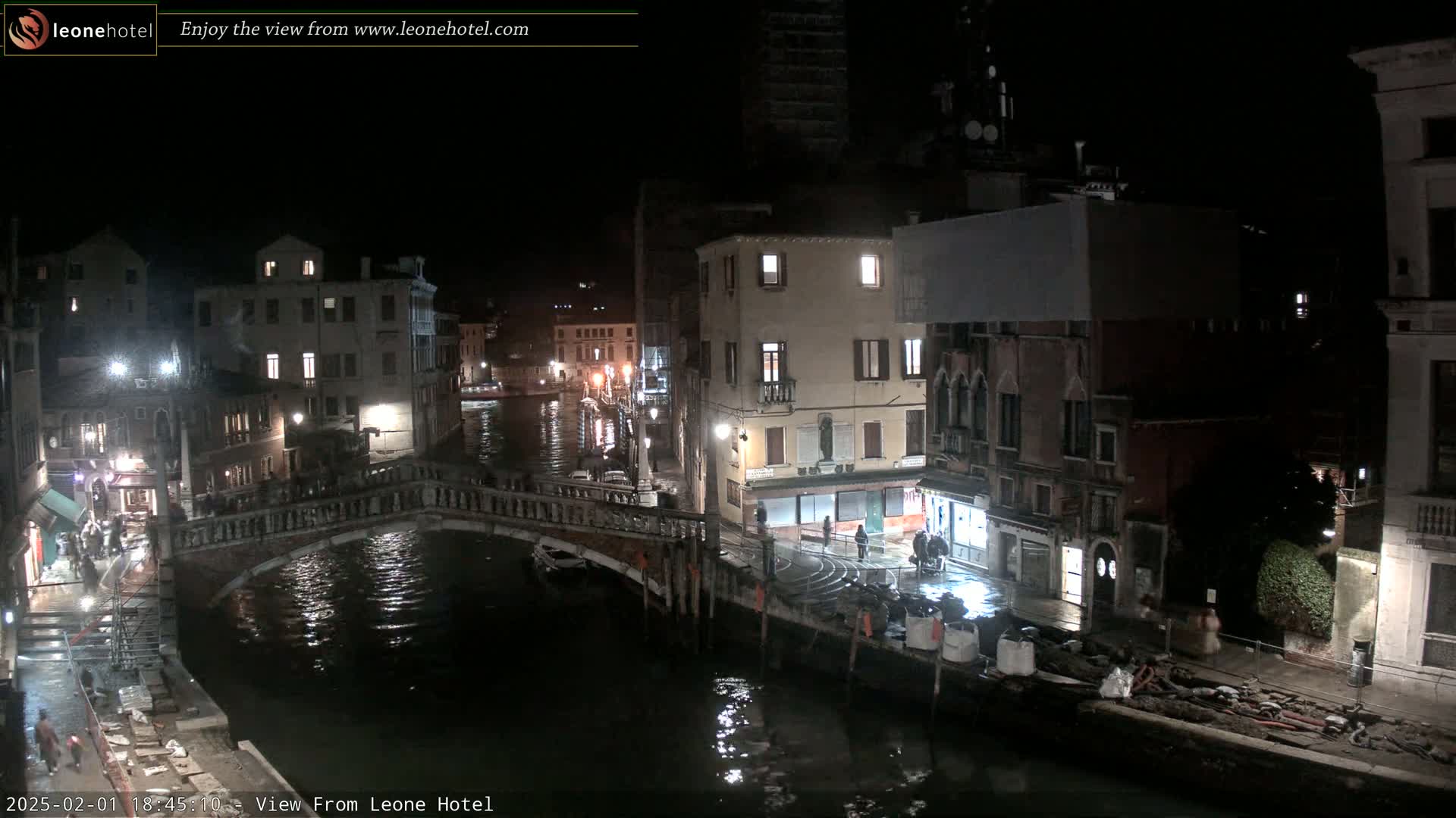 A nighttime view of a canal in Venice, showing a stone bridge, several buildings with lights on, and a dark, calm waterway.