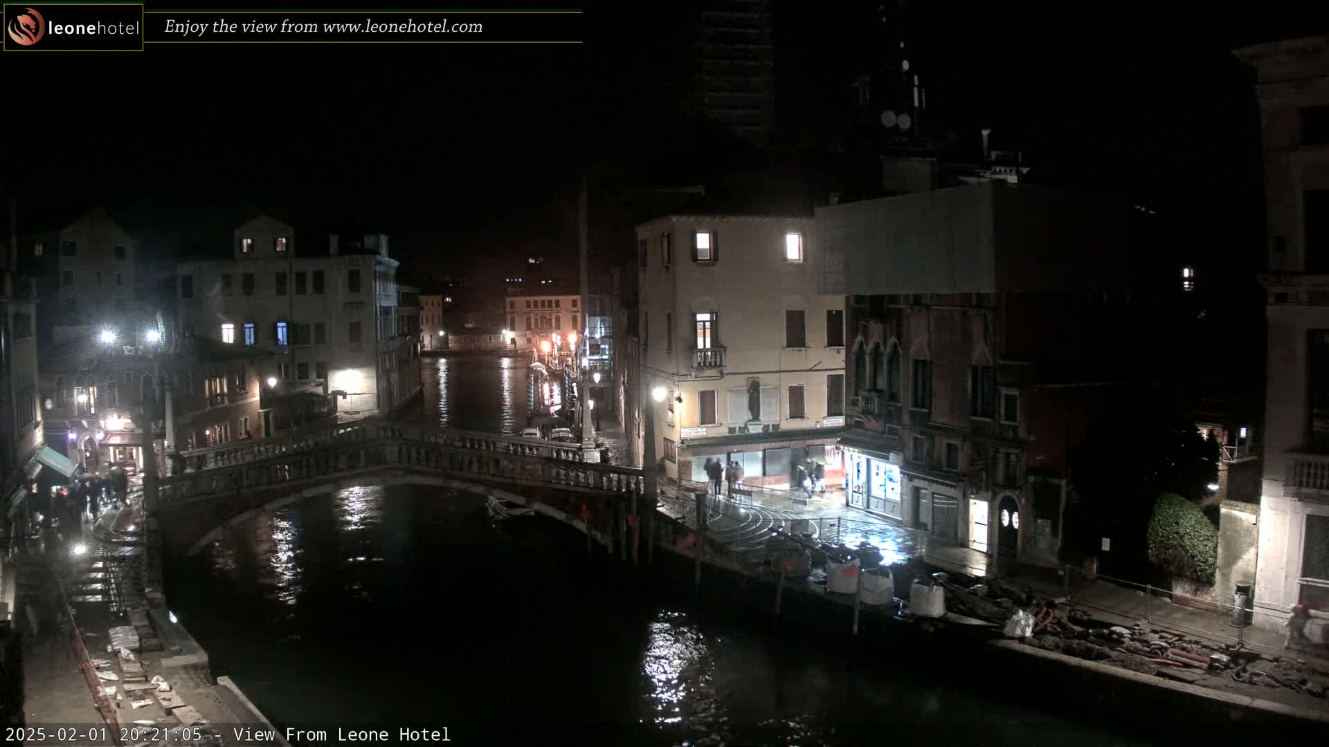 A nighttime view of a canal in Venice, Italy, shows a stone bridge spanning the water, with buildings lining both sides of the canal, under a clear, dark sky.