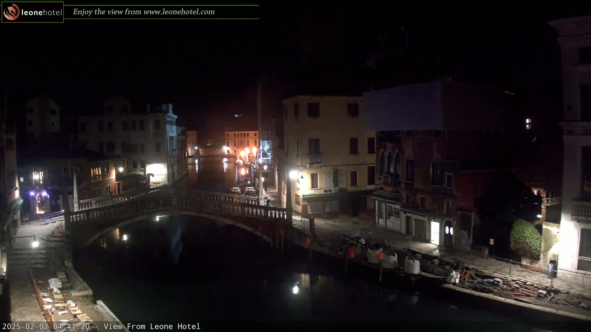 A nighttime view of a canal in Venice, Italy, shows several buildings along the waterway, a bridge spanning the canal, and some construction materials on the canal bank.