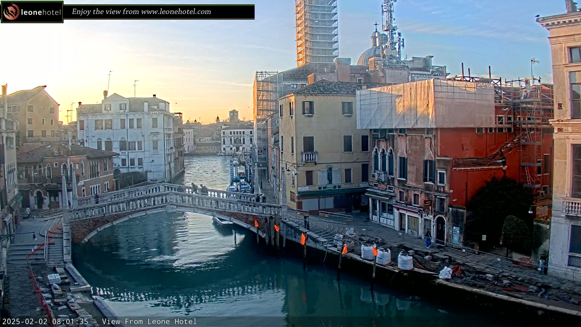 A bridge arches over a canal in Venice, Italy, at sunrise, with several buildings under construction or renovation along the waterway.