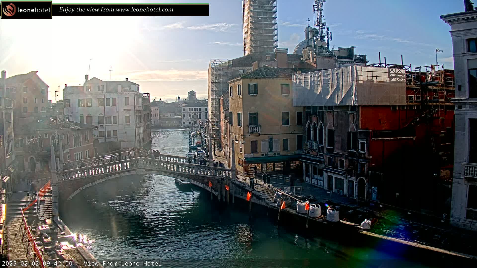 A sunny day in Venice shows a canal with a bridge, buildings along its banks, some under construction, and a few people walking.