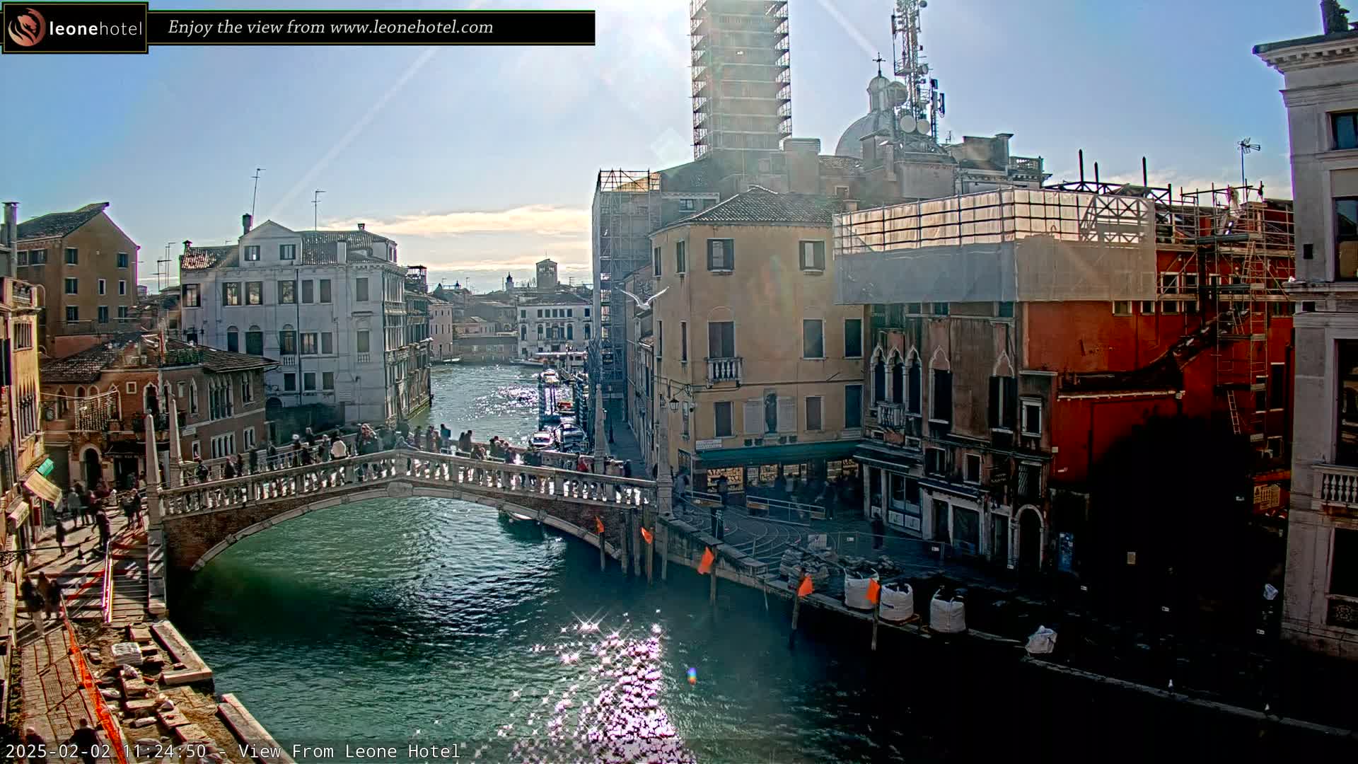A sunny day in Venice shows a canal with a bridge filled with people, surrounded by historic buildings, some of which are under construction.