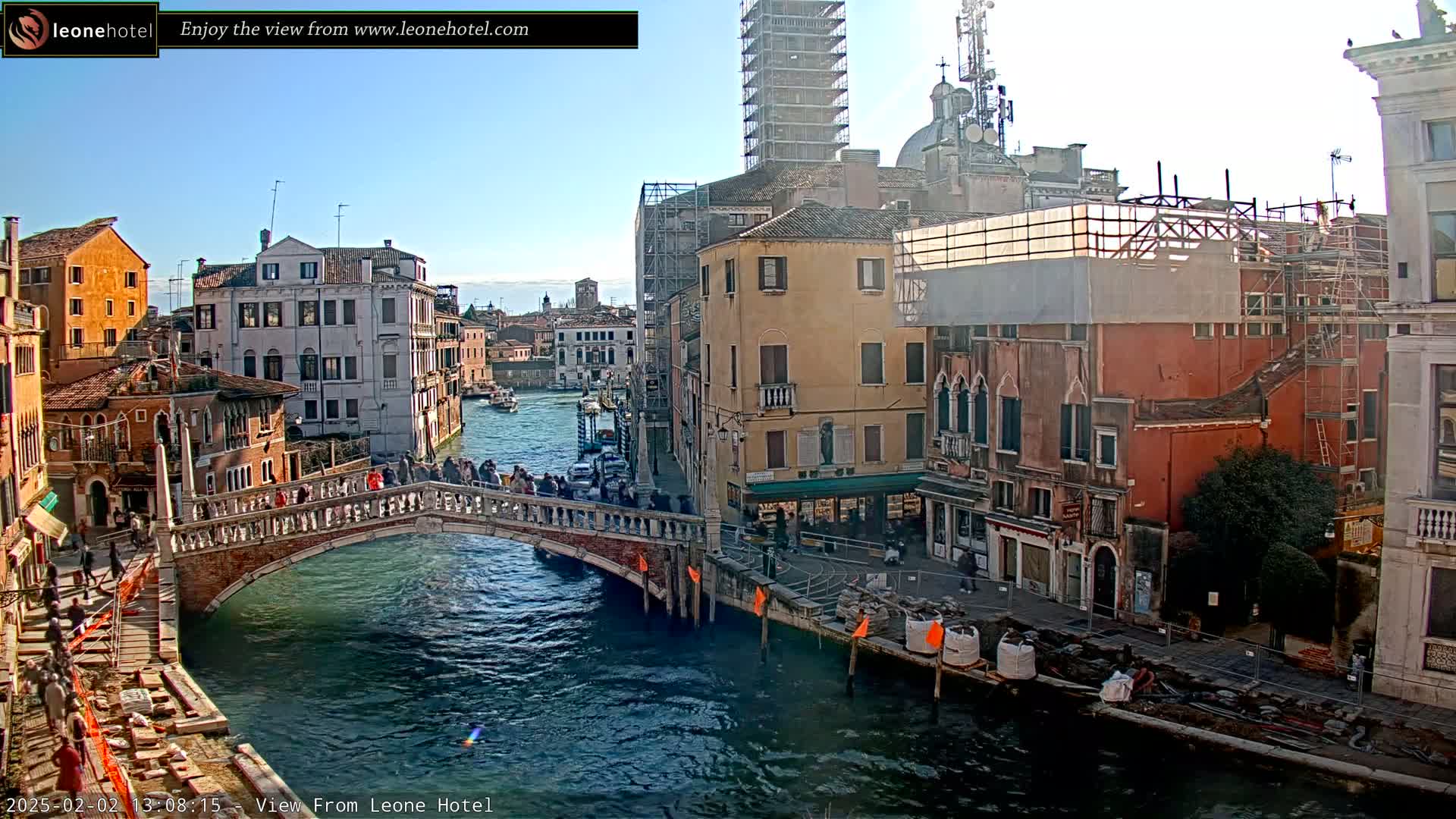 A sunny day in Venice shows a canal with a stone bridge crowded with people, colorful buildings lining the waterway, and construction scaffolding visible on some of the structures.