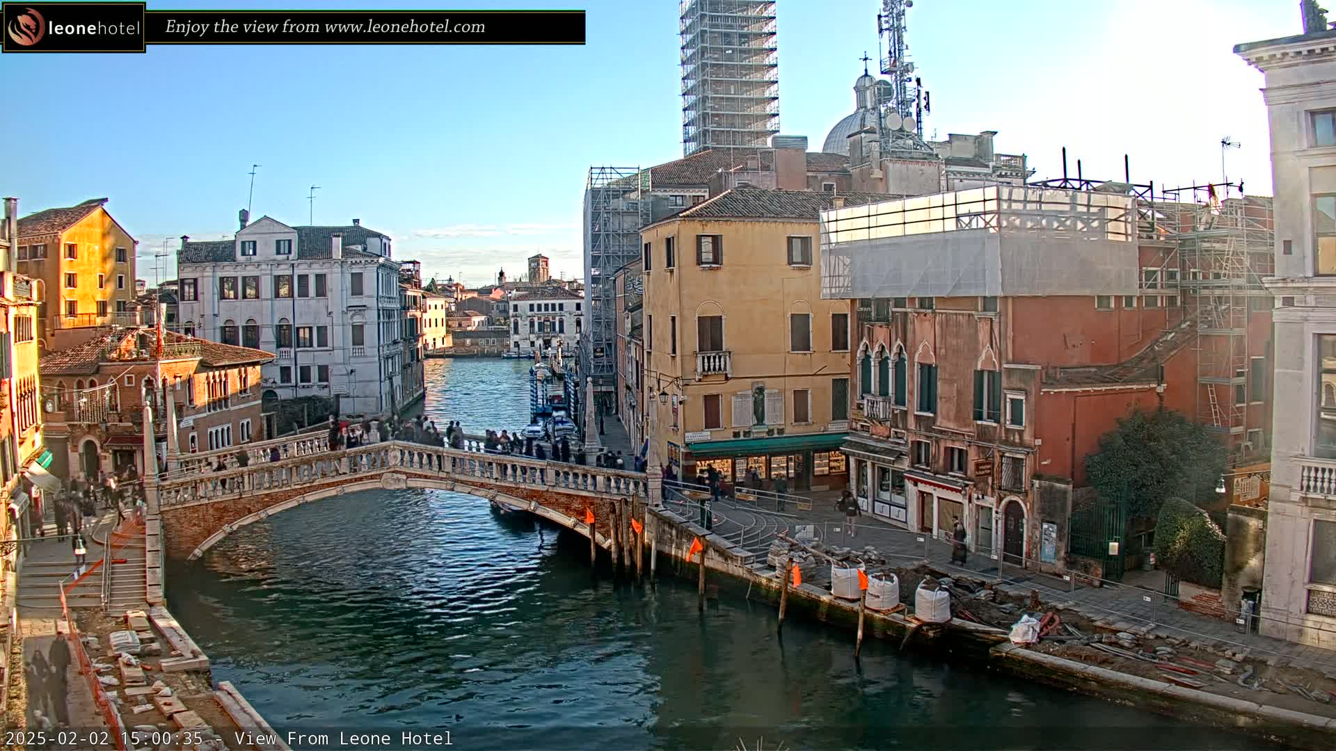 A canal in Venice, Italy, under a clear blue sky, shows a stone bridge with people walking across it, and several buildings, some under construction.
