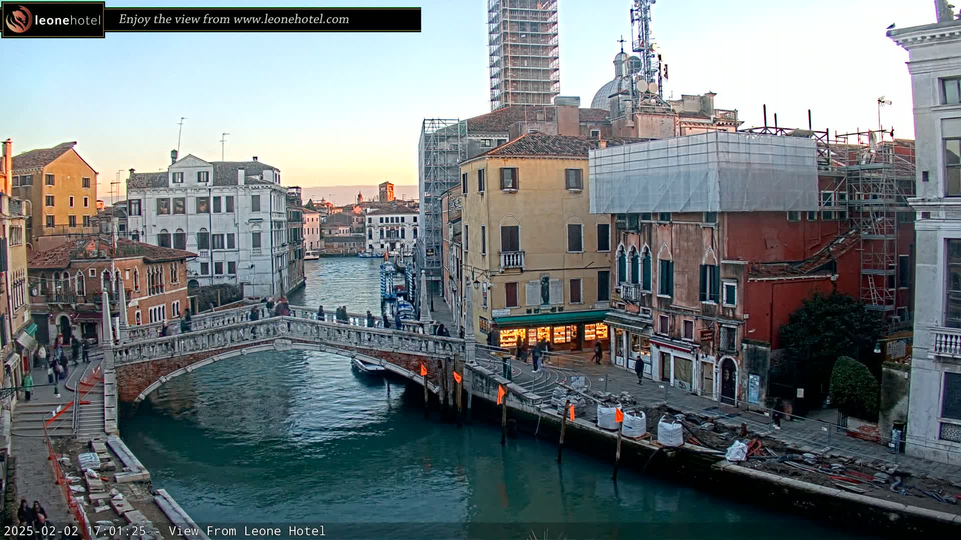 A stone bridge spans a canal in Venice, Italy, under a twilight sky, with several buildings along the waterway undergoing construction or renovations.